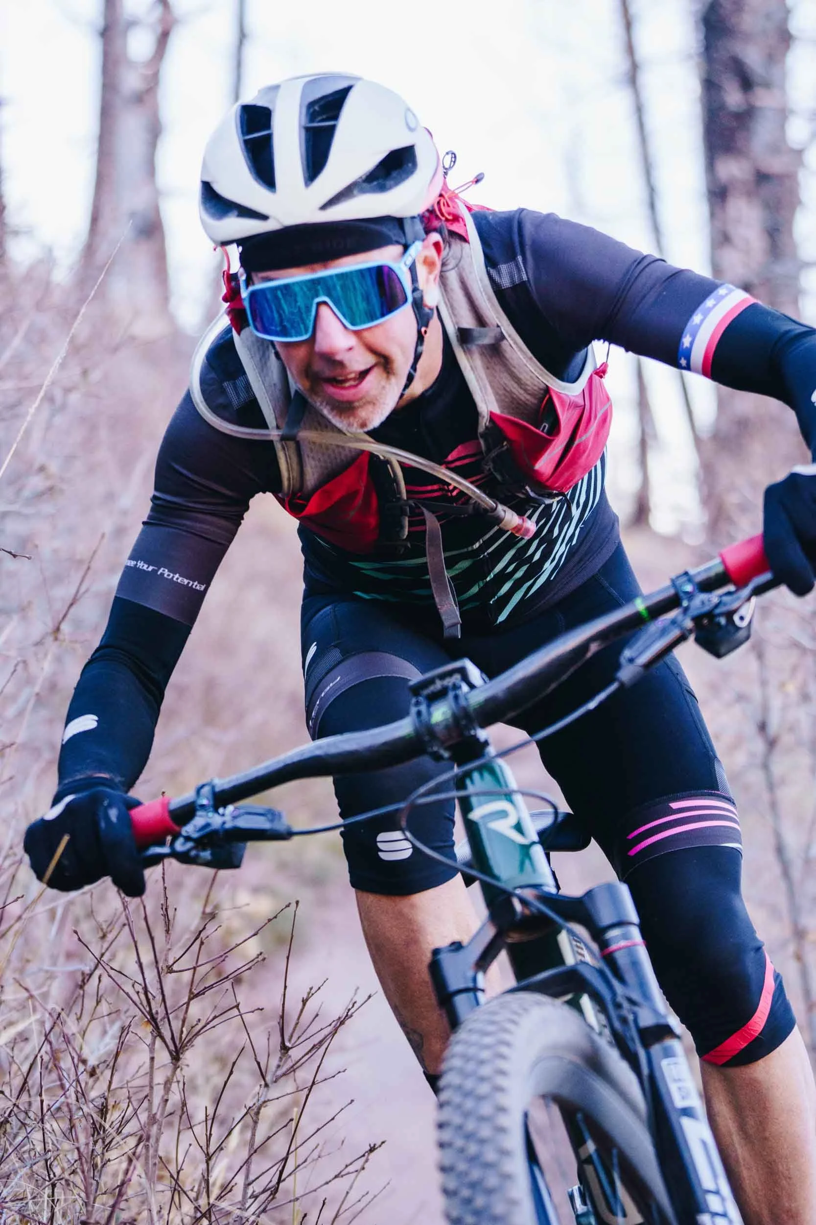 Nick mountain biking on a dirt trail through a forest, wearing a helmet, sunglasses, and athletic gear.