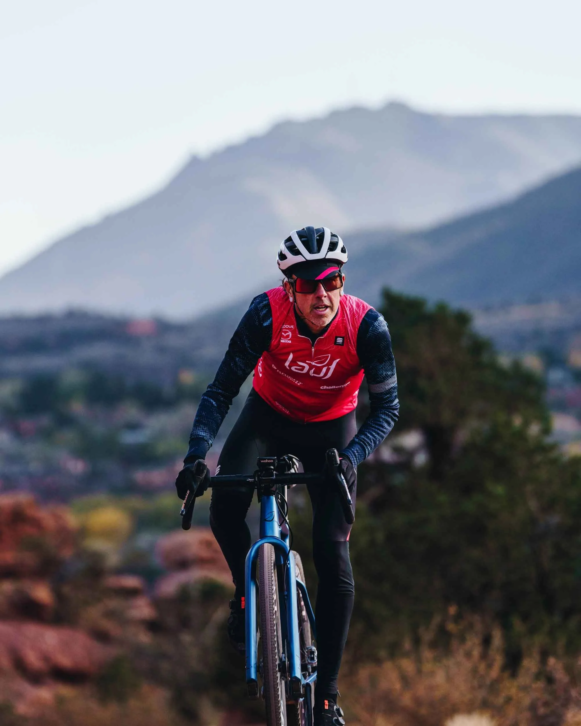 Nick Gould professional cyclist wearing a helmet, sunglasses, and a red and black athletic outfit rides mountain biking on a trail with mountainous terrain in the background.