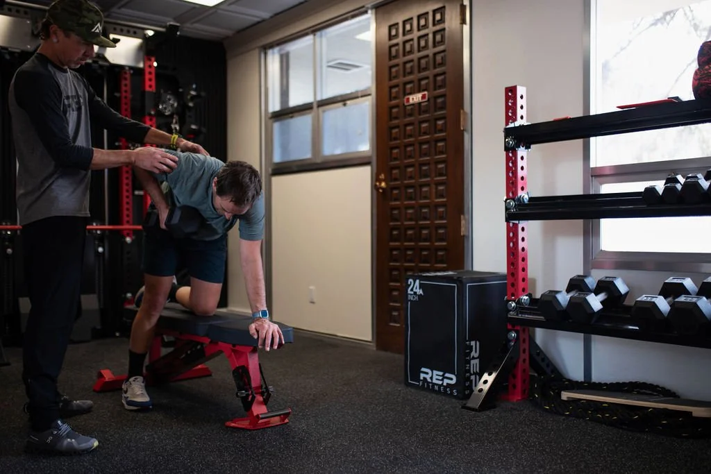 Nick Gould certified personal trainer assists a man performing a push-up on a bench in a gym, with weightlifting equipment visible in the background.