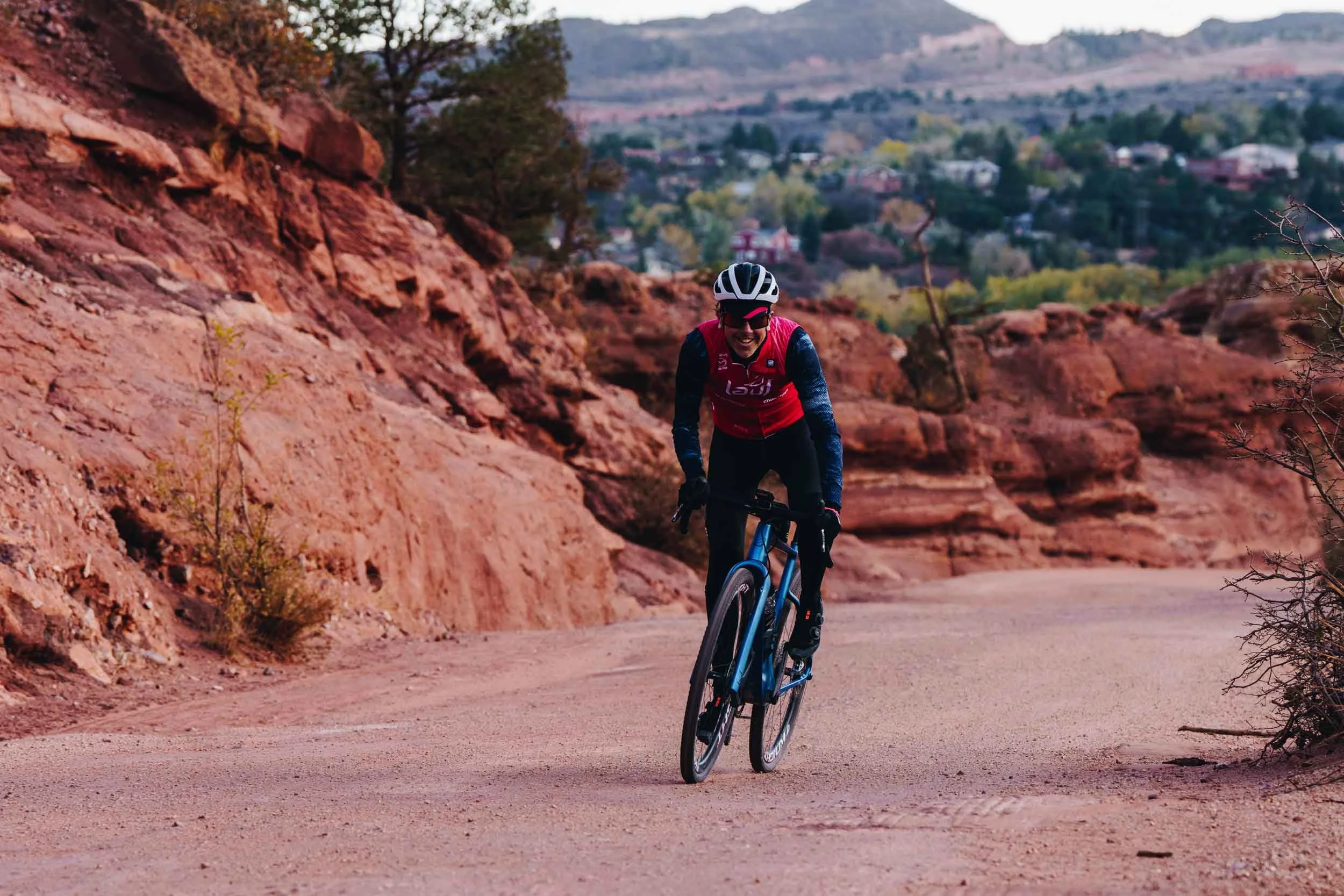 Nick mountain biking on a dirt trail through red rock landscape with a town visible in the background.