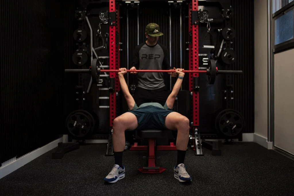 Client lying on a bench doing a bench press with a barbell, assisted by a personal trainer Nick Gould in his private PurEnergy Perfrmance Durango Colorado gym.