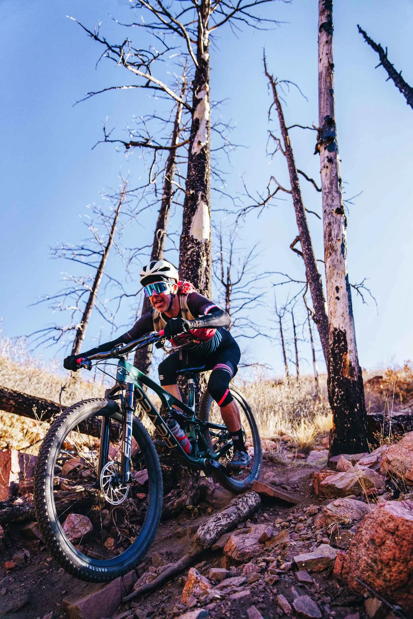 Nick Gould, a professional mountain biker wearing a white helmet, sunglasses, black and pink cycling gear, descending a rocky trail in a forest with leafless trees and a clear blue sky.