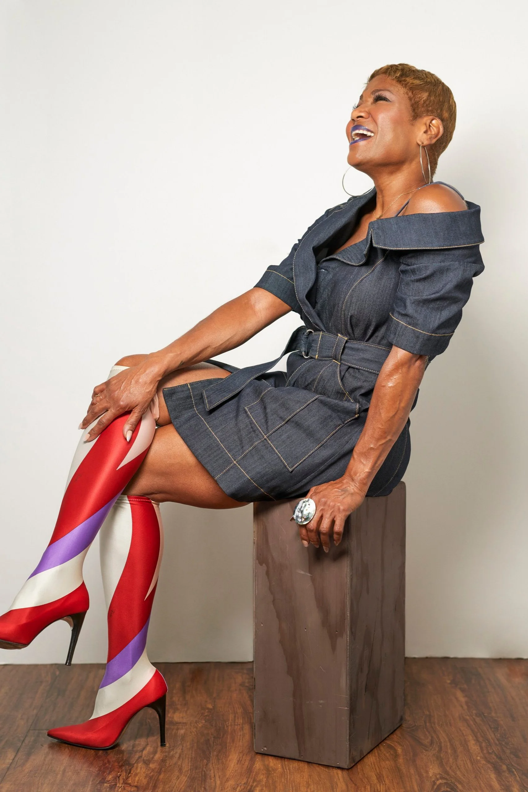 A woman wearing a denim dress and colorful striped high heels is sitting on a wooden block, laughing joyfully against a plain light background.
