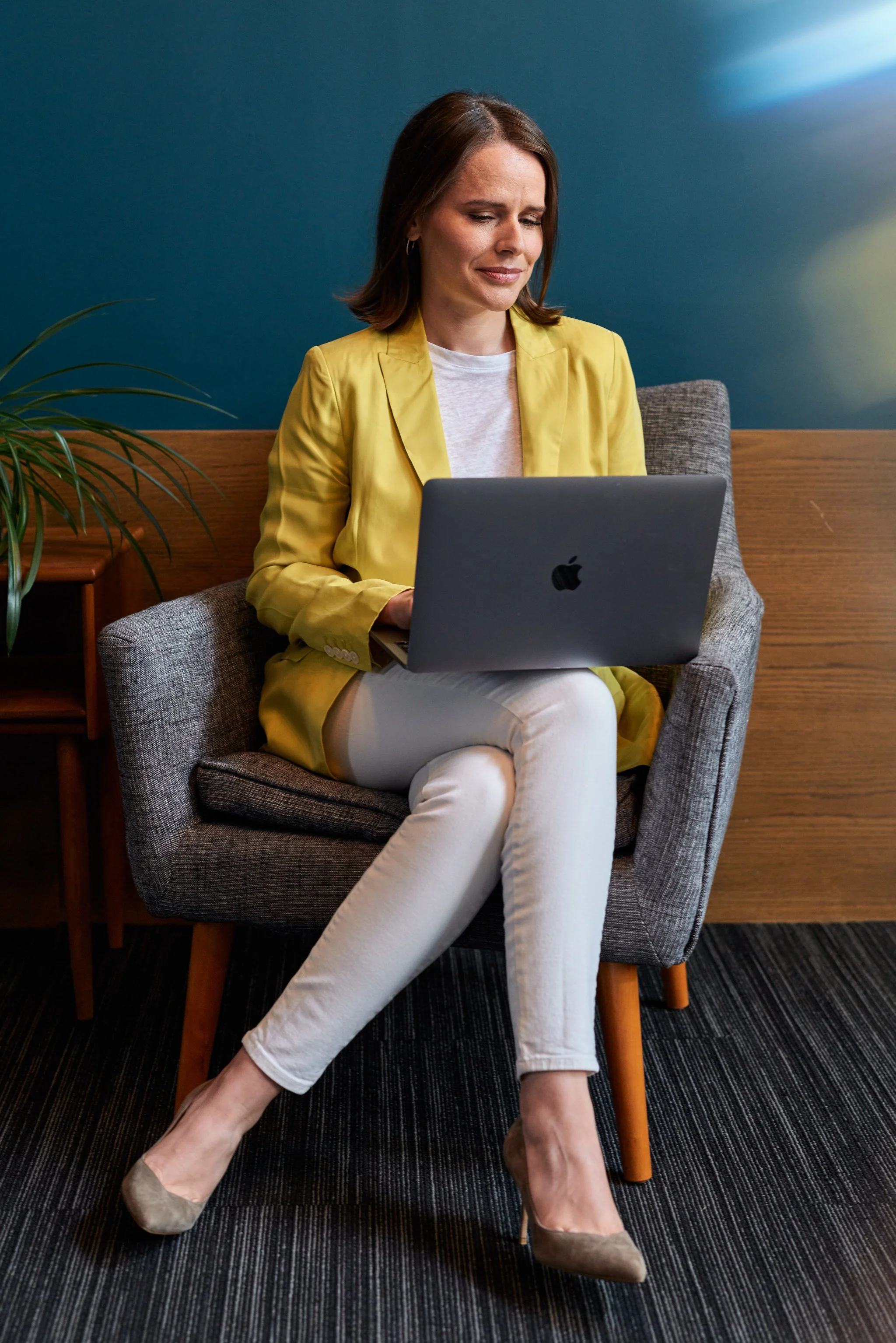 A woman with shoulder-length brown hair, wearing a yellow blazer, white pants, and beige heels, sitting on a gray armchair with a gray laptop on her lap, looking at the screen with a slight frown. She is in a room with a teal wall, a wooden side table with a plant, and a black carpet.