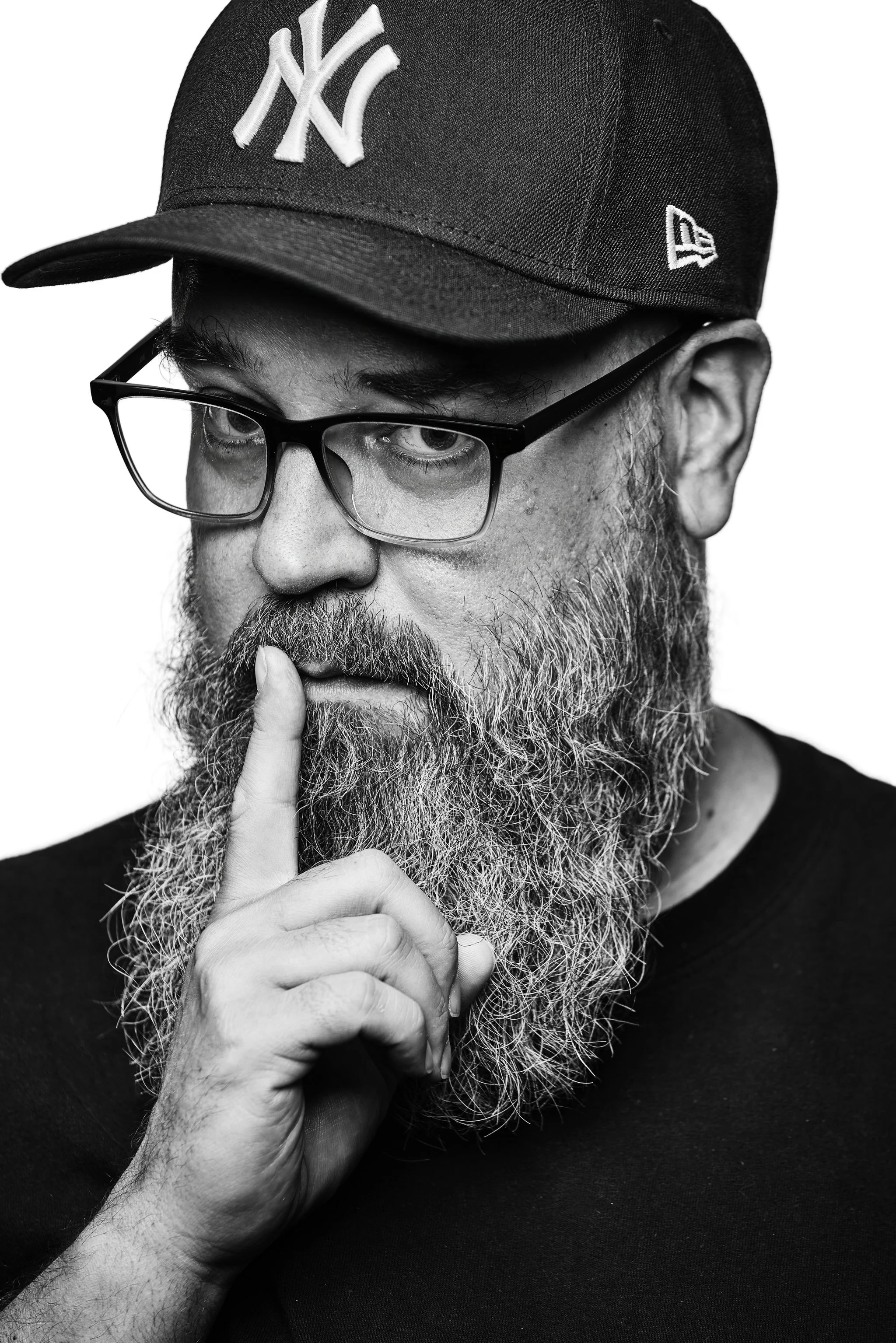 A black-and-white portrait of a man with glasses, a beard, and a mustache wearing a New York Yankees cap and touching his lips with his finger.