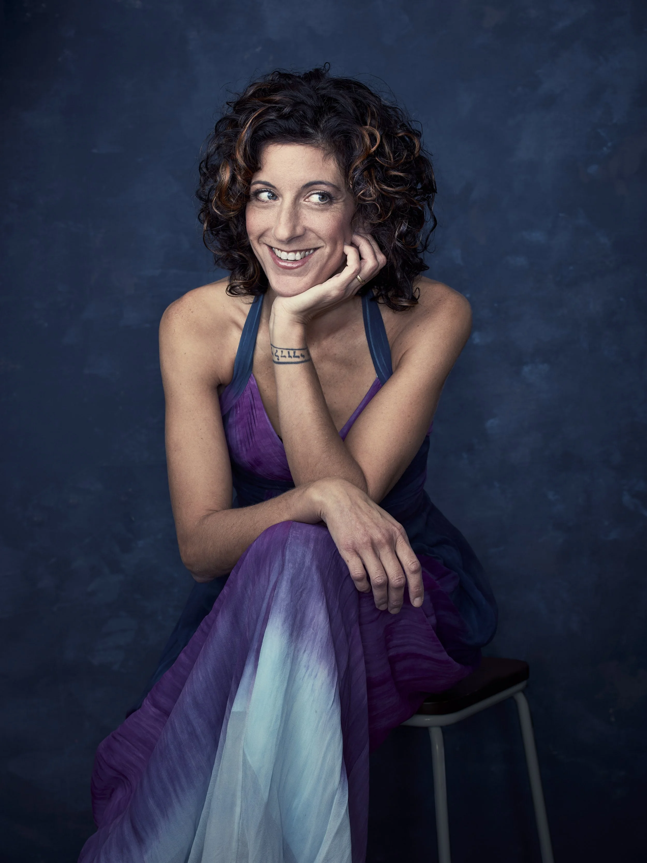 A woman with curly dark hair, smiling and resting her chin on her hand, sitting on a stool against a dark blue background.