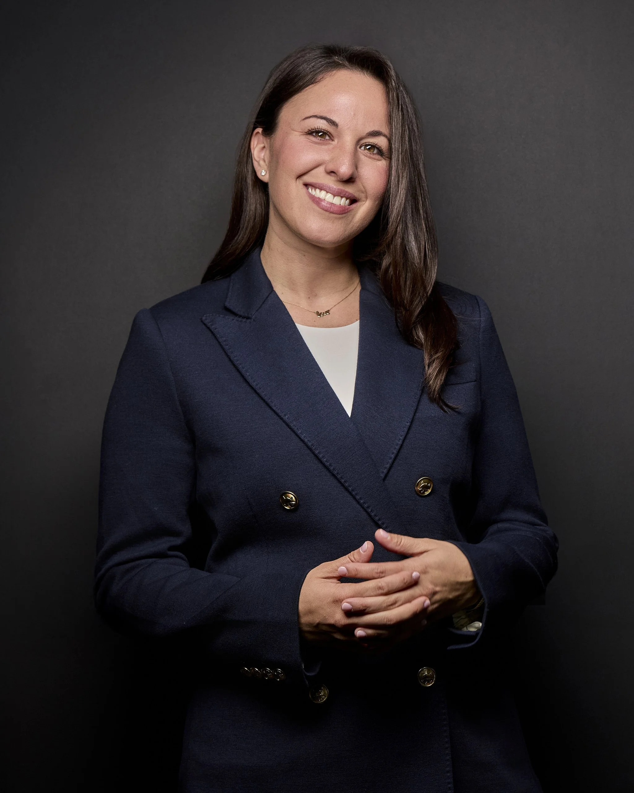 A woman with long brown hair, wearing a navy blazer over a white top, smiling and standing against a dark background.
