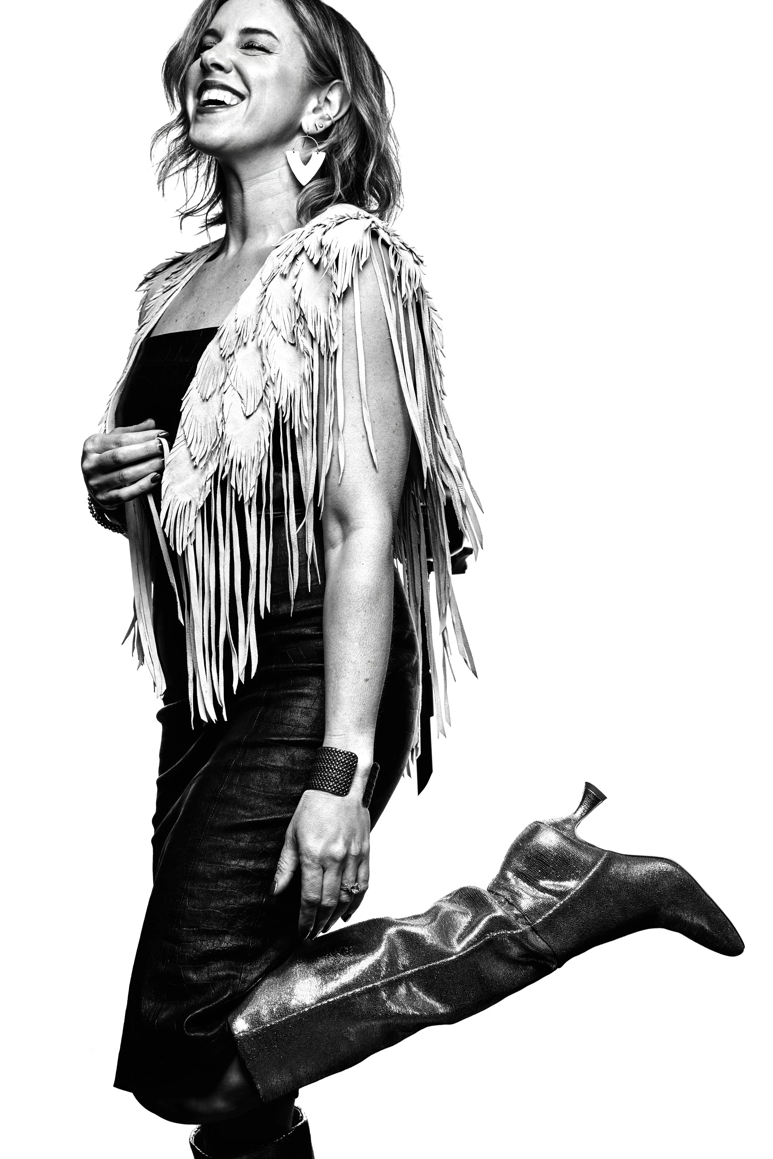 Black and white photo of a woman smiling, wearing a fringed jacket, black top, leather pants, and high boots, with one leg bent and raised.