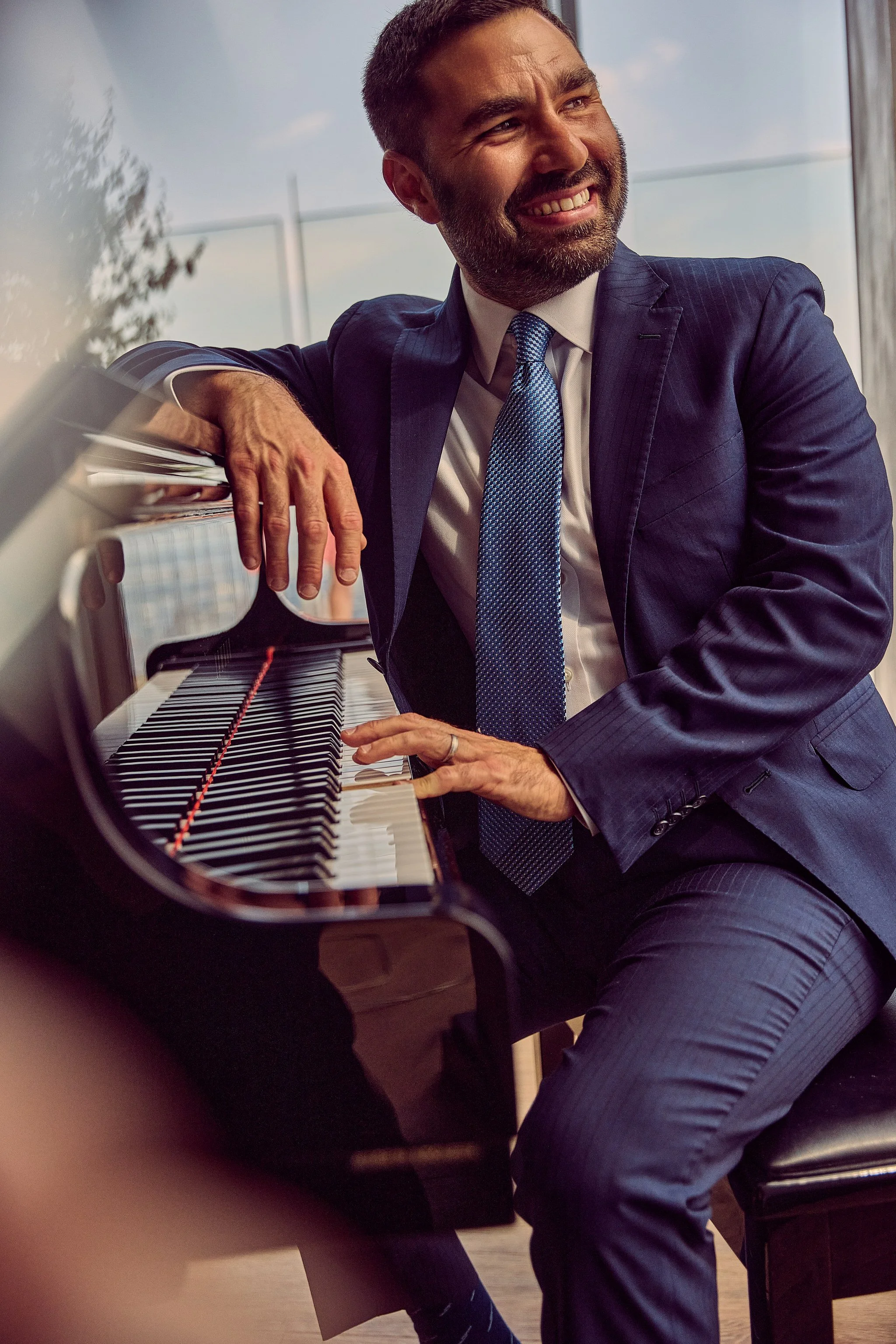 A man dressed in a suit and tie playing a grand piano. He is smiling and looking up with a window in the background.