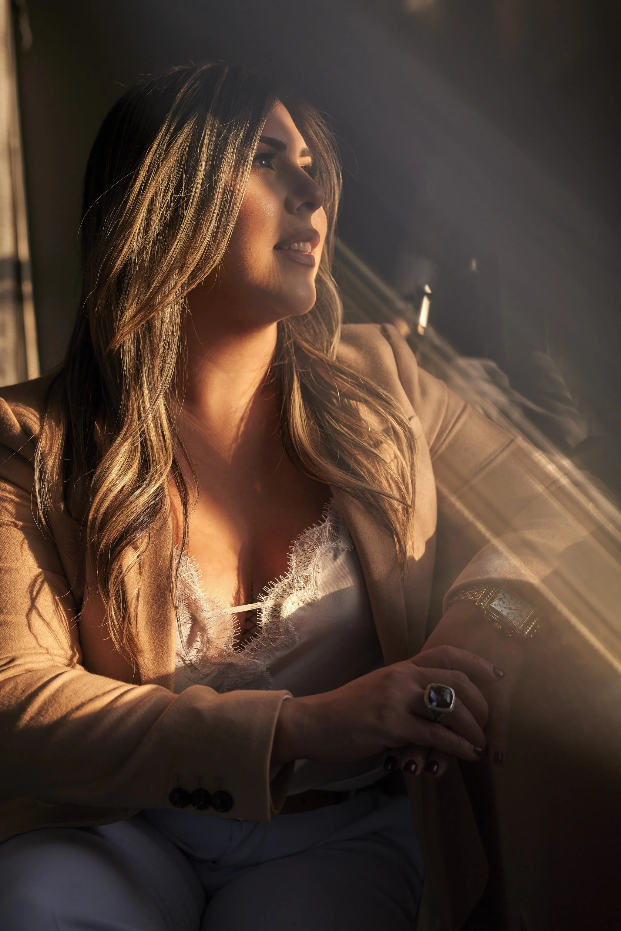 A woman with long hair sitting by a window, wearing a beige blazer, white lace top, and jewelry, looking out the window with sunlight on her face.