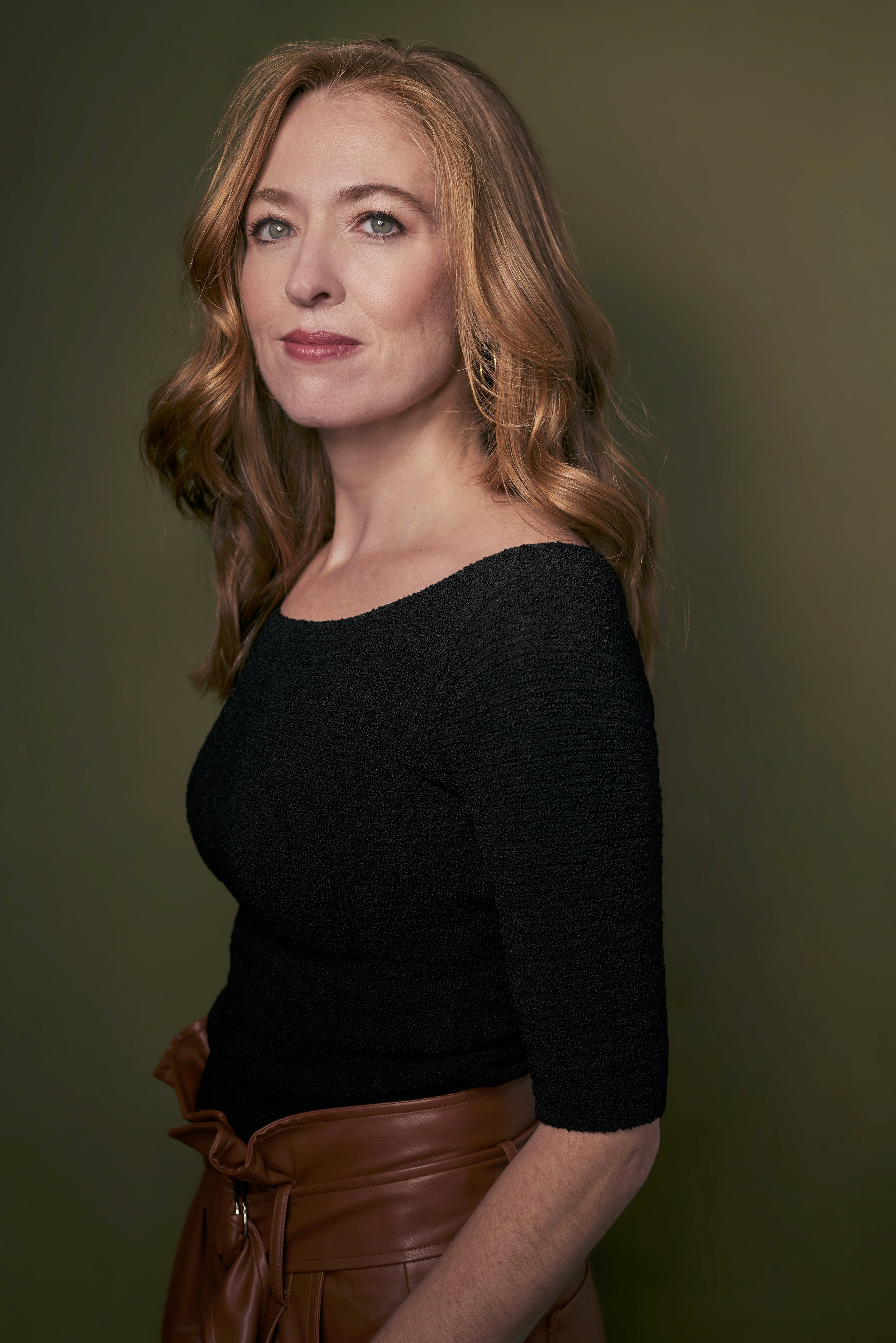 A woman with shoulder-length red hair and blue eyes wearing a black top and brown pants with a large bow, standing against a green background.