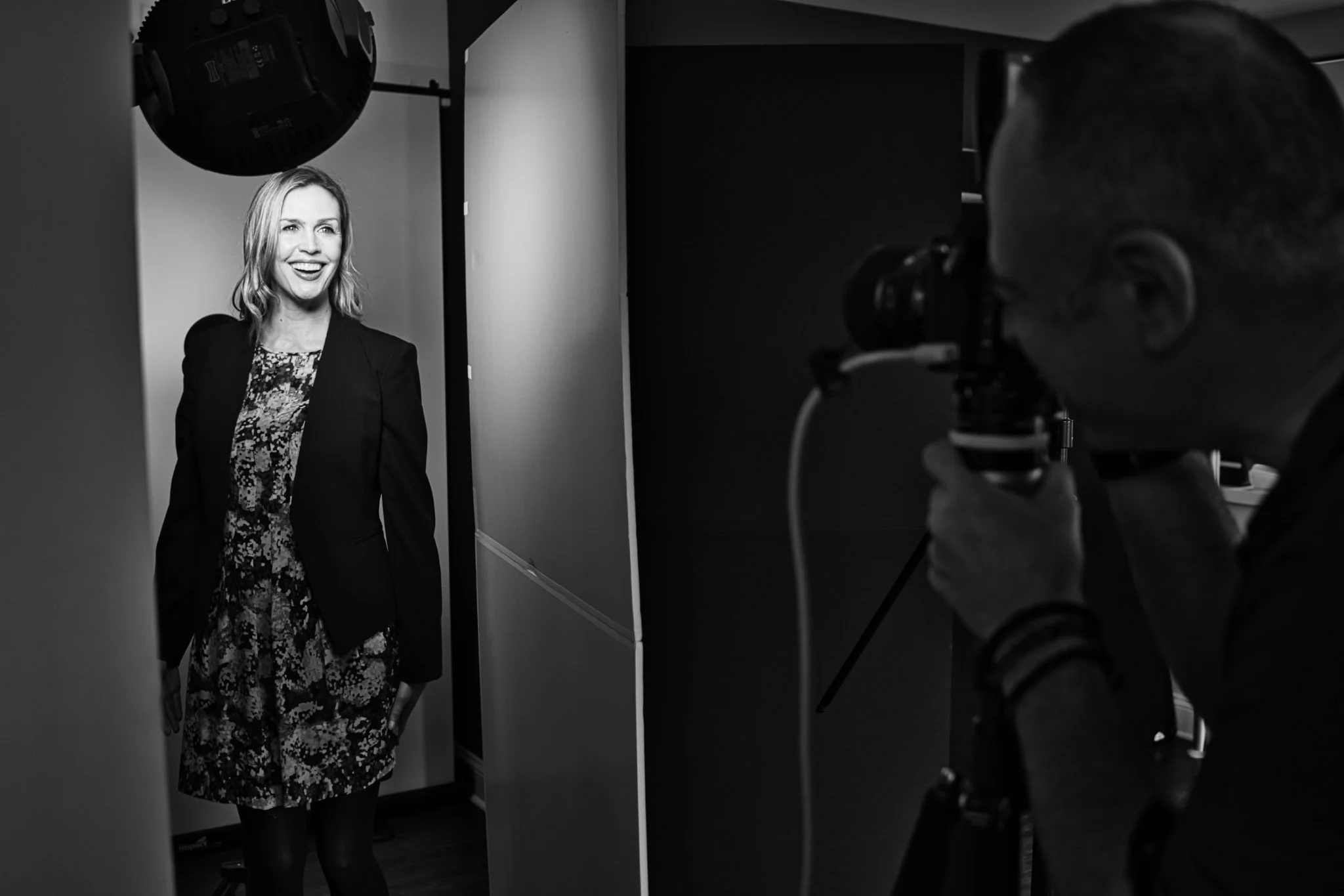 A woman stands in front of a camera, smiling and looking to her left, during a photoshoot in a studio with a black-and-white filter.