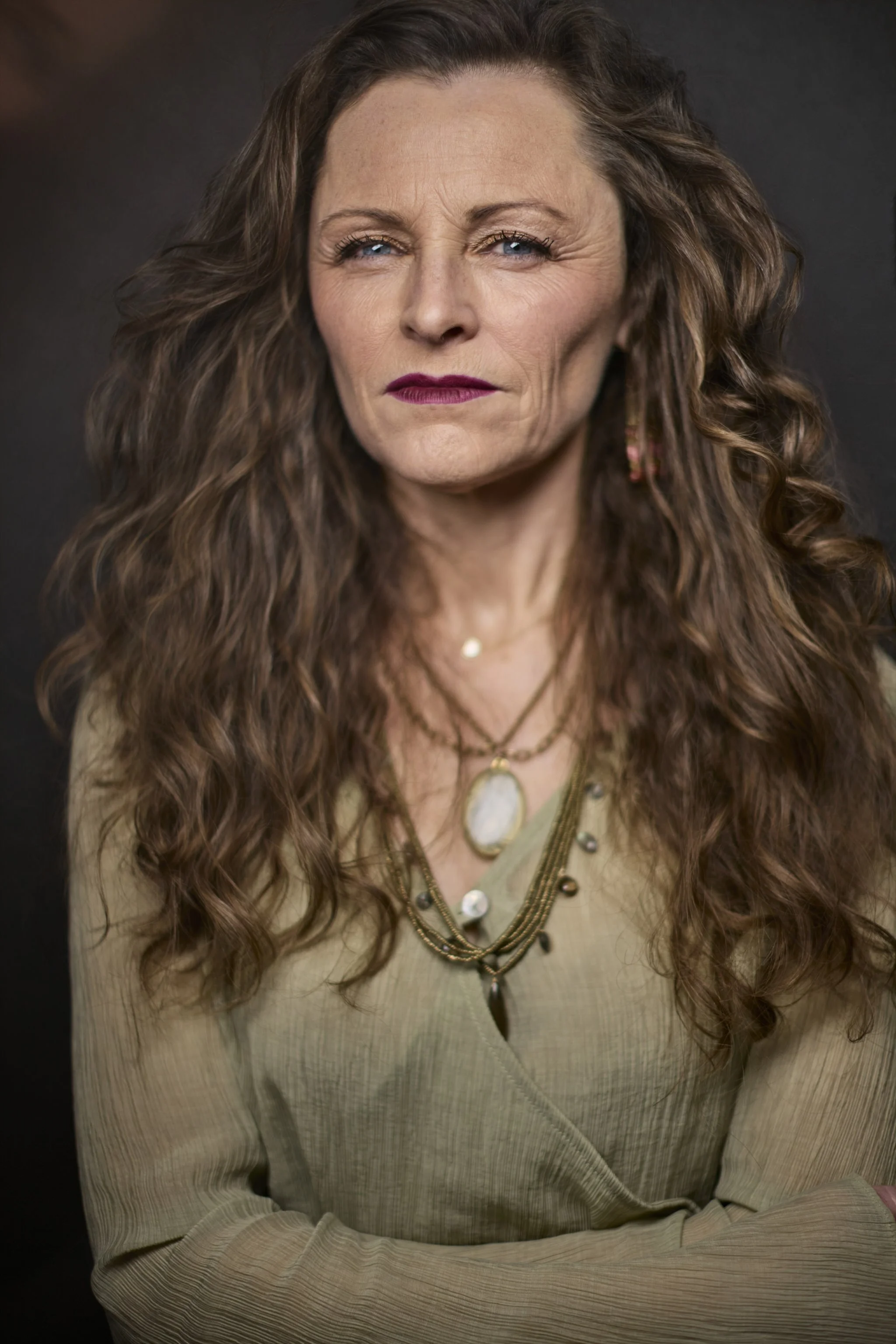 A confident middle-aged woman with long, wavy brown hair, wearing a beige outfit and layered necklaces, looking directly at the camera with her arms crossed.