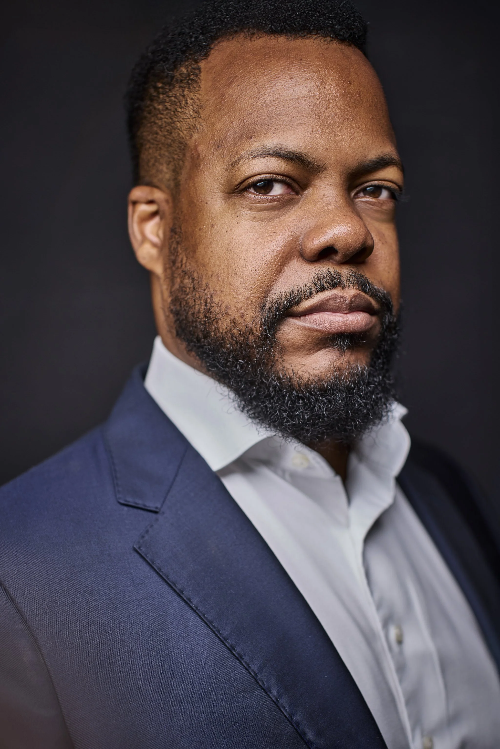 Close-up portrait of an African American man with short curly black hair, a full beard, wearing a navy suit jacket over a white dress shirt, against a dark background.