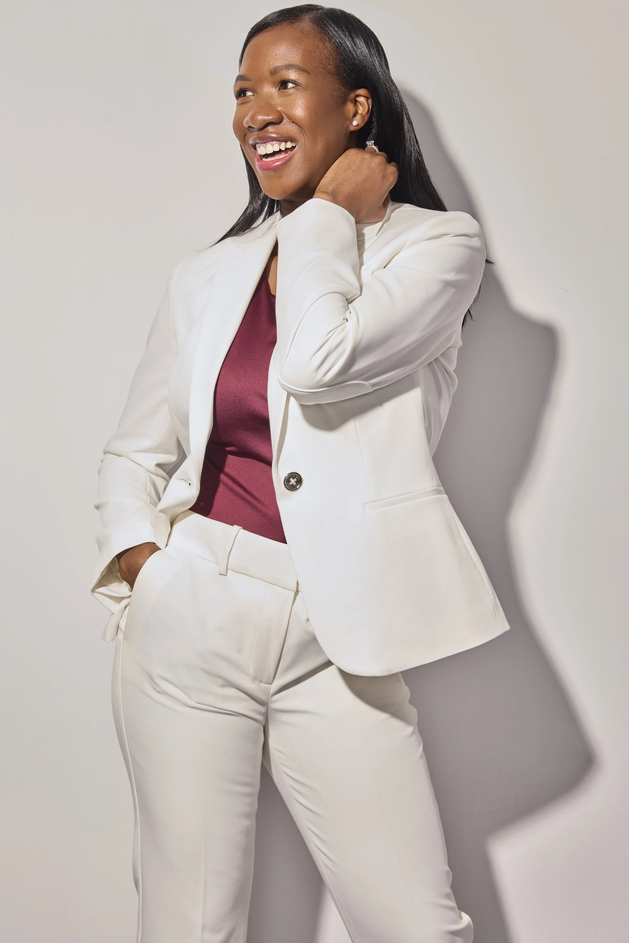 A woman smiling, wearing a white blazer and pants with a maroon top, standing against a plain white background.