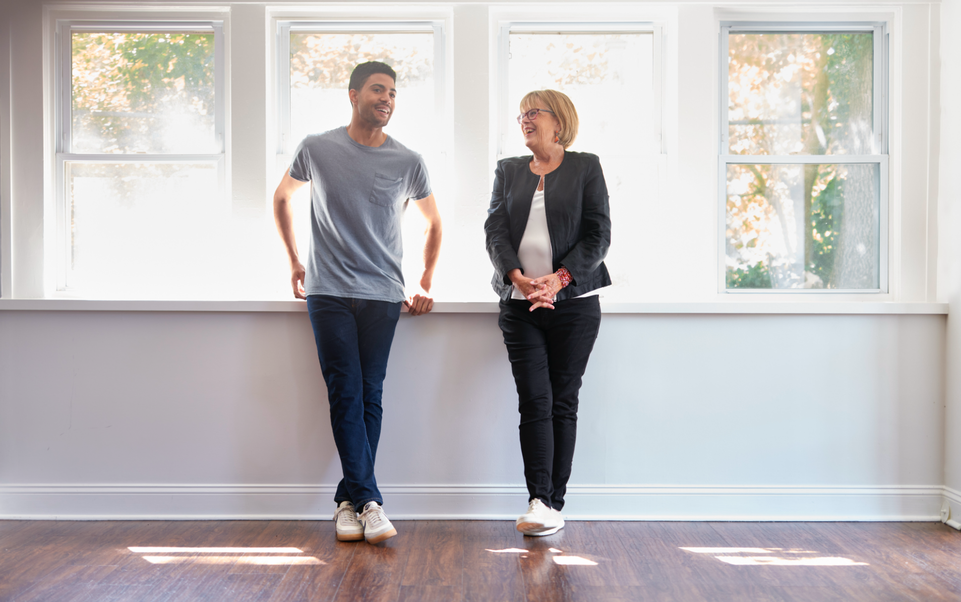 A young man and an older woman are standing indoors near large windows with trees outside, smiling and conversing.