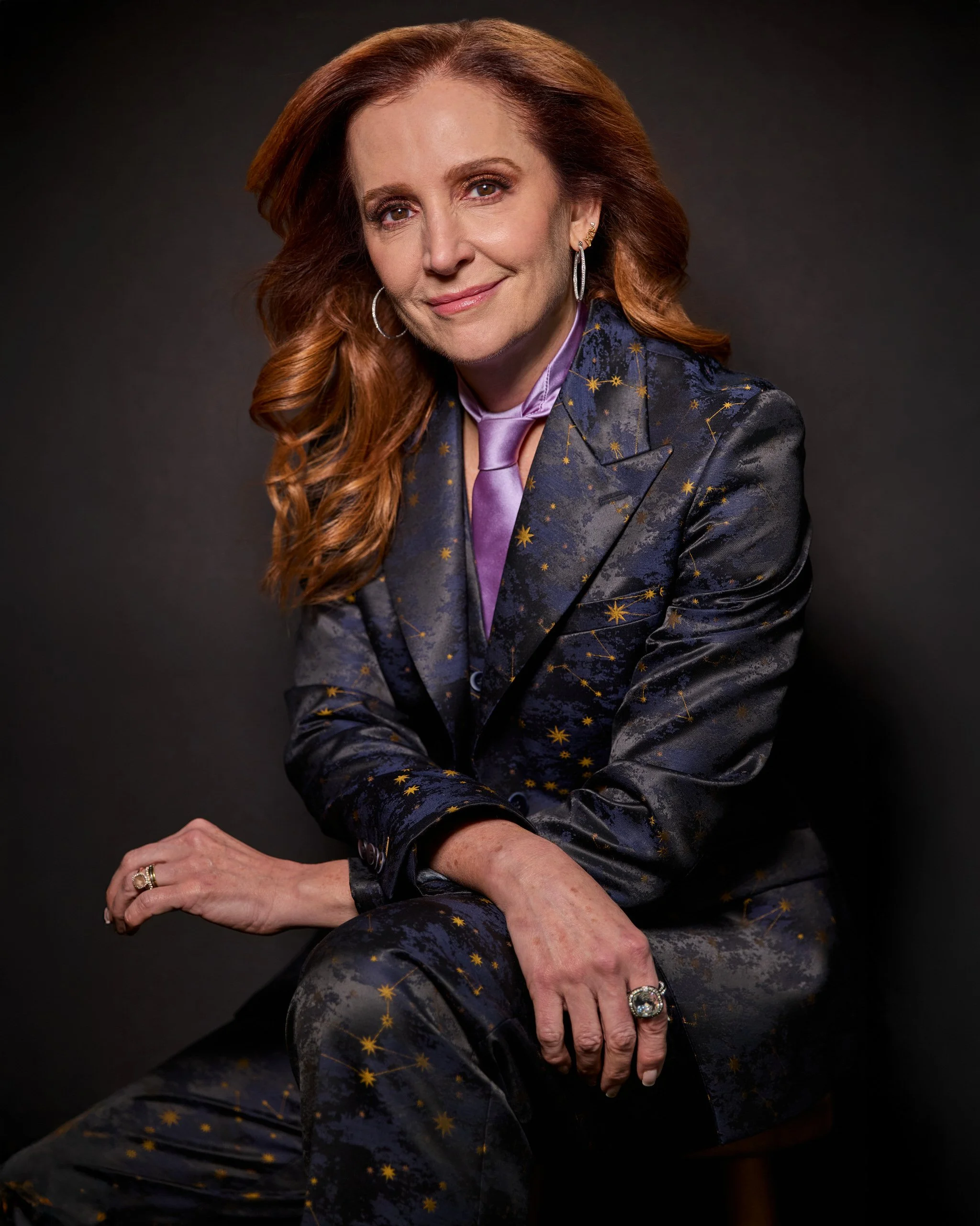 A woman with long red hair wearing a dark starry patterned suit, a purple silk blouse, and jewelry, posing against a dark background.