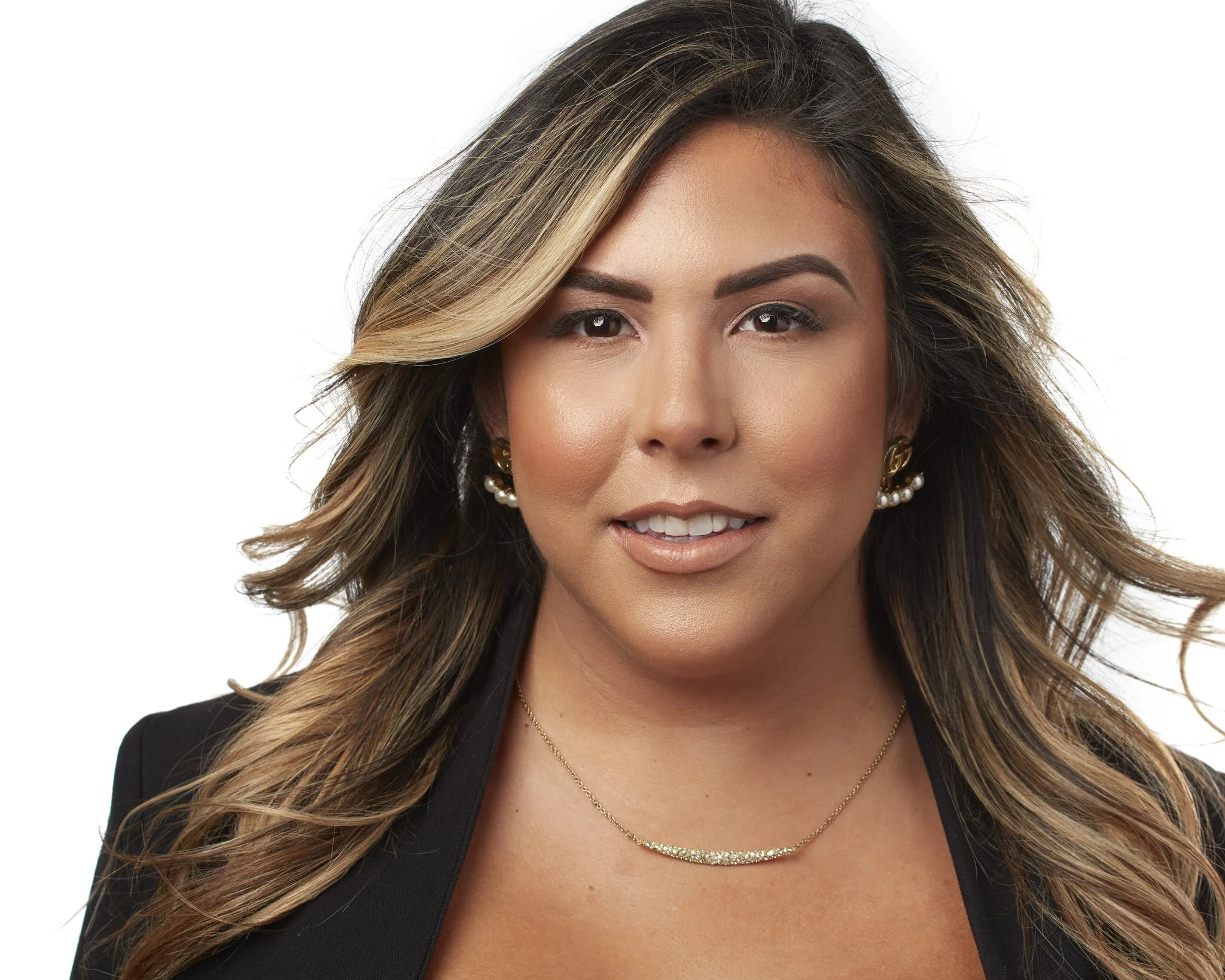 Portrait of a woman with long, wavy hair, wearing gold earrings, a necklace, and a black blazer, smiling slightly, against a white background.