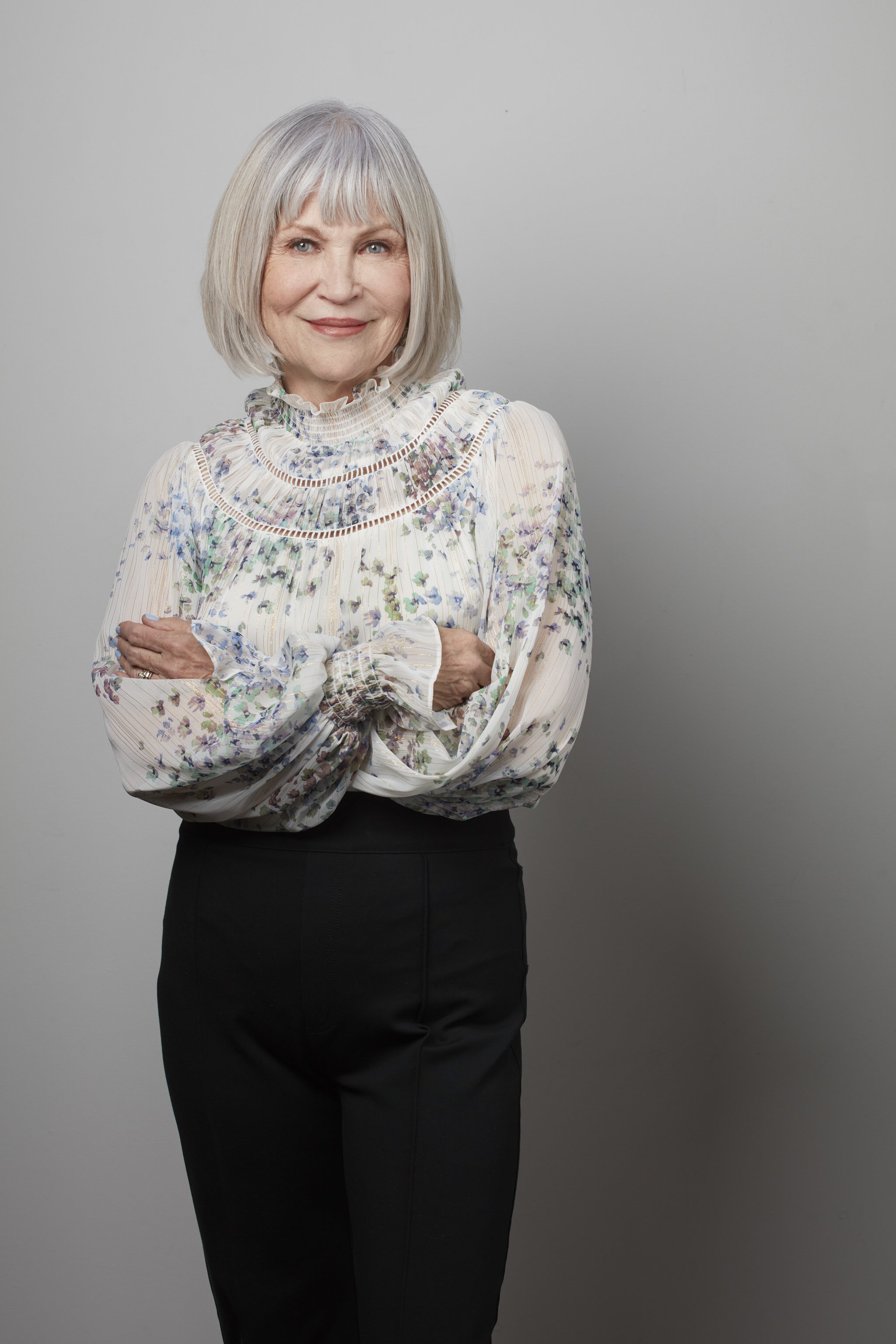 Older woman with gray hair wearing a floral blouse and black pants, standing with arms crossed, smiling, against a plain gray background.