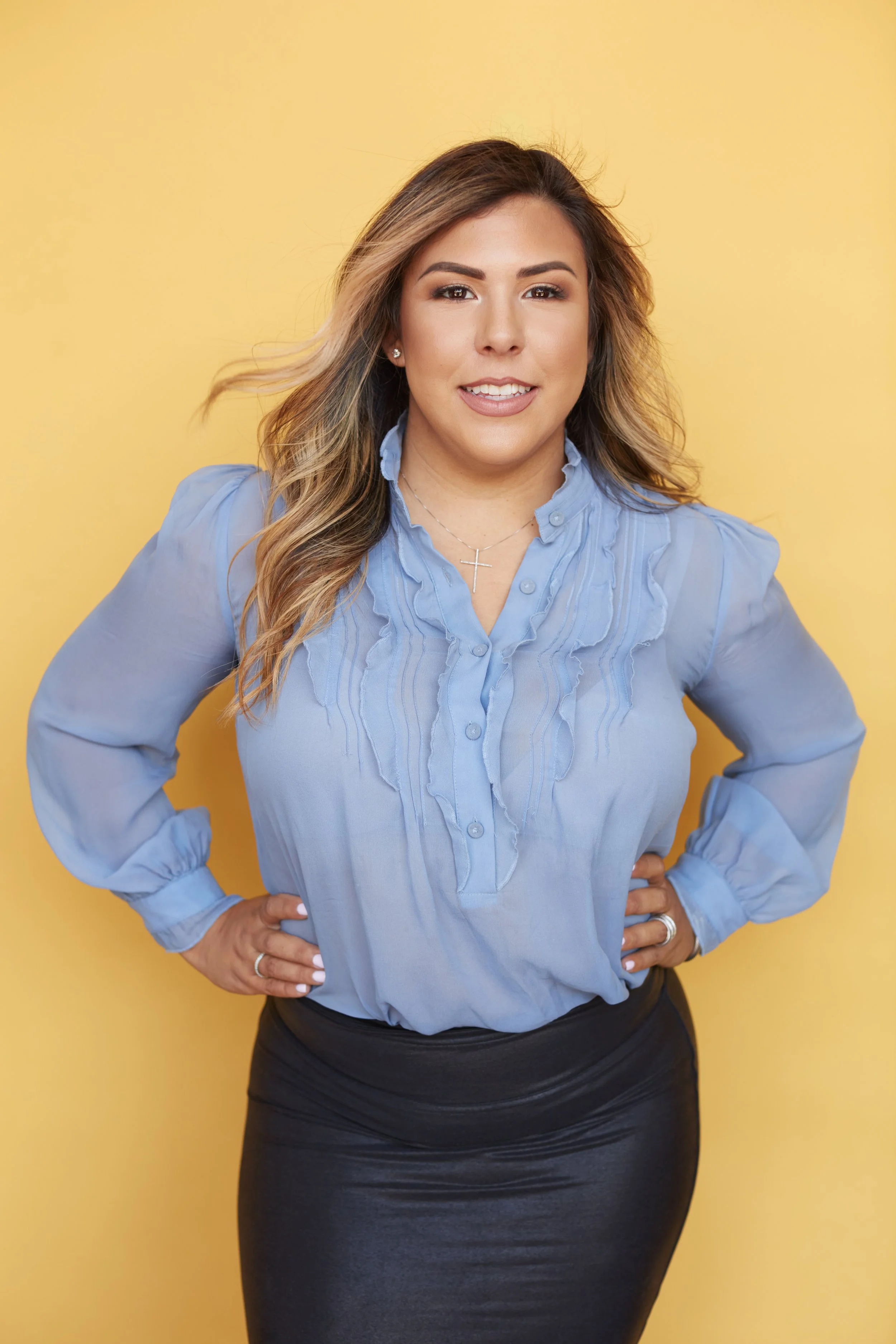 Woman with blonde hair wearing a light blue blouse and a black skirt, standing against a yellow background, smiling with hands on her hips.