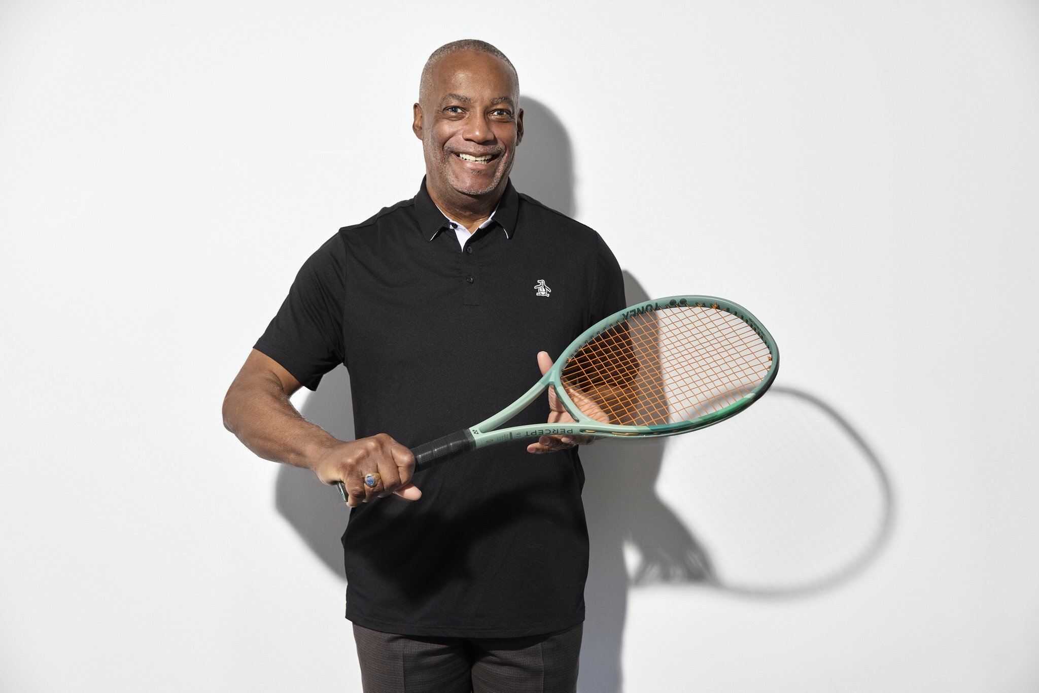A middle-aged man with short gray hair, wearing a black polo shirt, holding a tennis racket, smiling, standing against a white background.