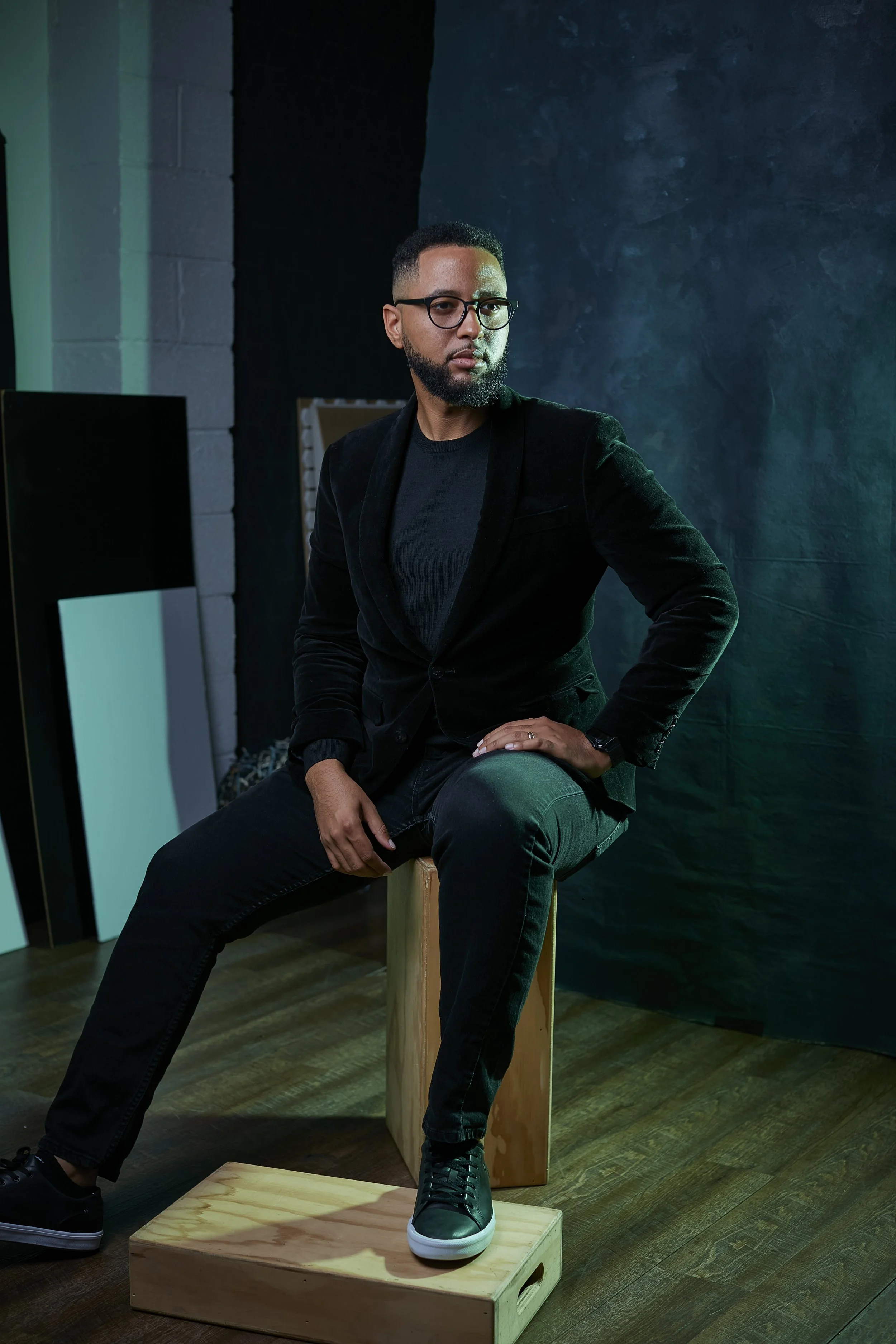 A man with glasses and a beard, dressed in black, sitting on a wooden box in a dimly lit studio with dark walls, looking to his right.