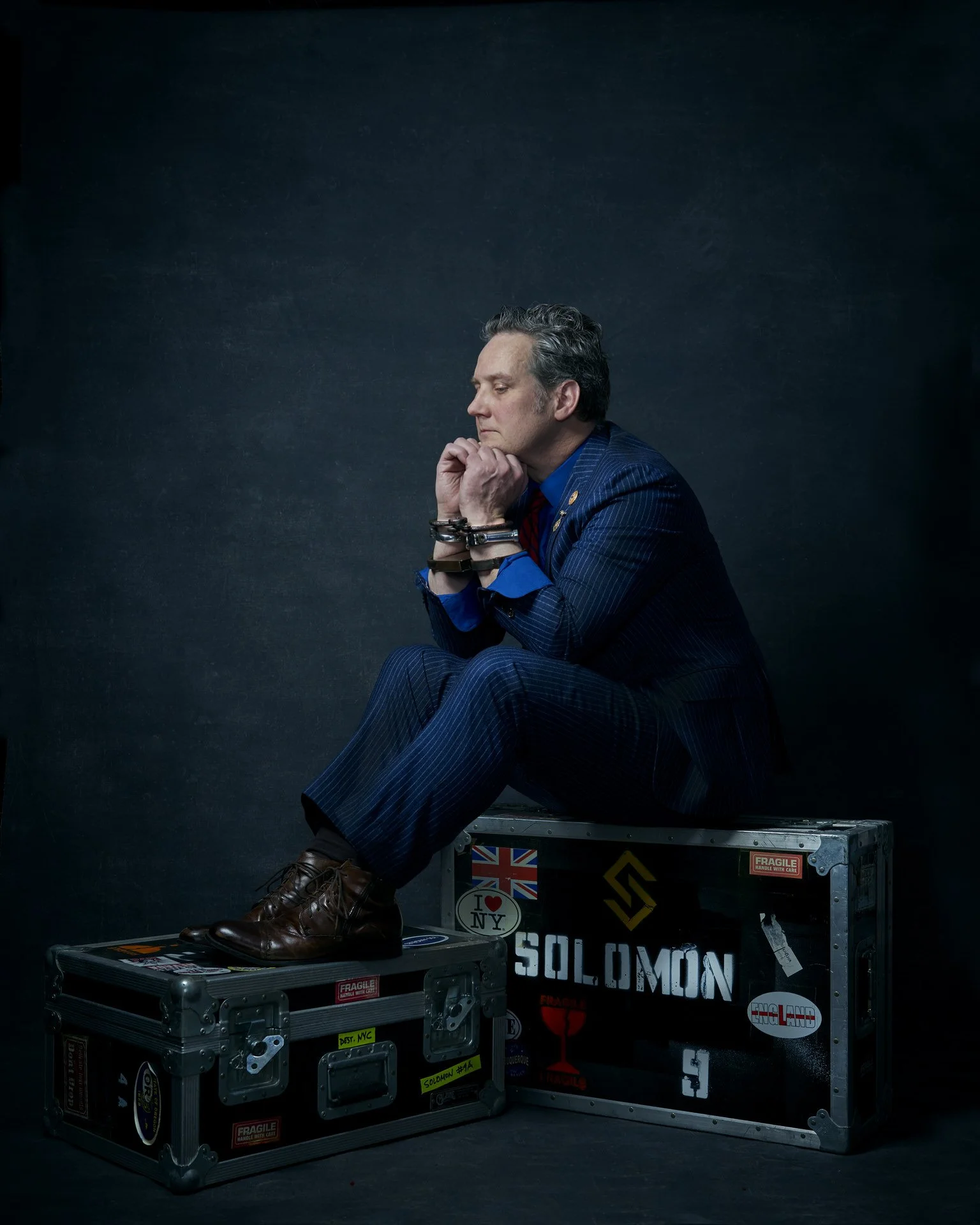 A man in a blue striped suit sits on a flight case with his hands clasped near his chin, looking contemplative against a dark background. The case has stickers including a UK flag, a red wine glass with 'I love NY,' and the word 'SOLOMON.'