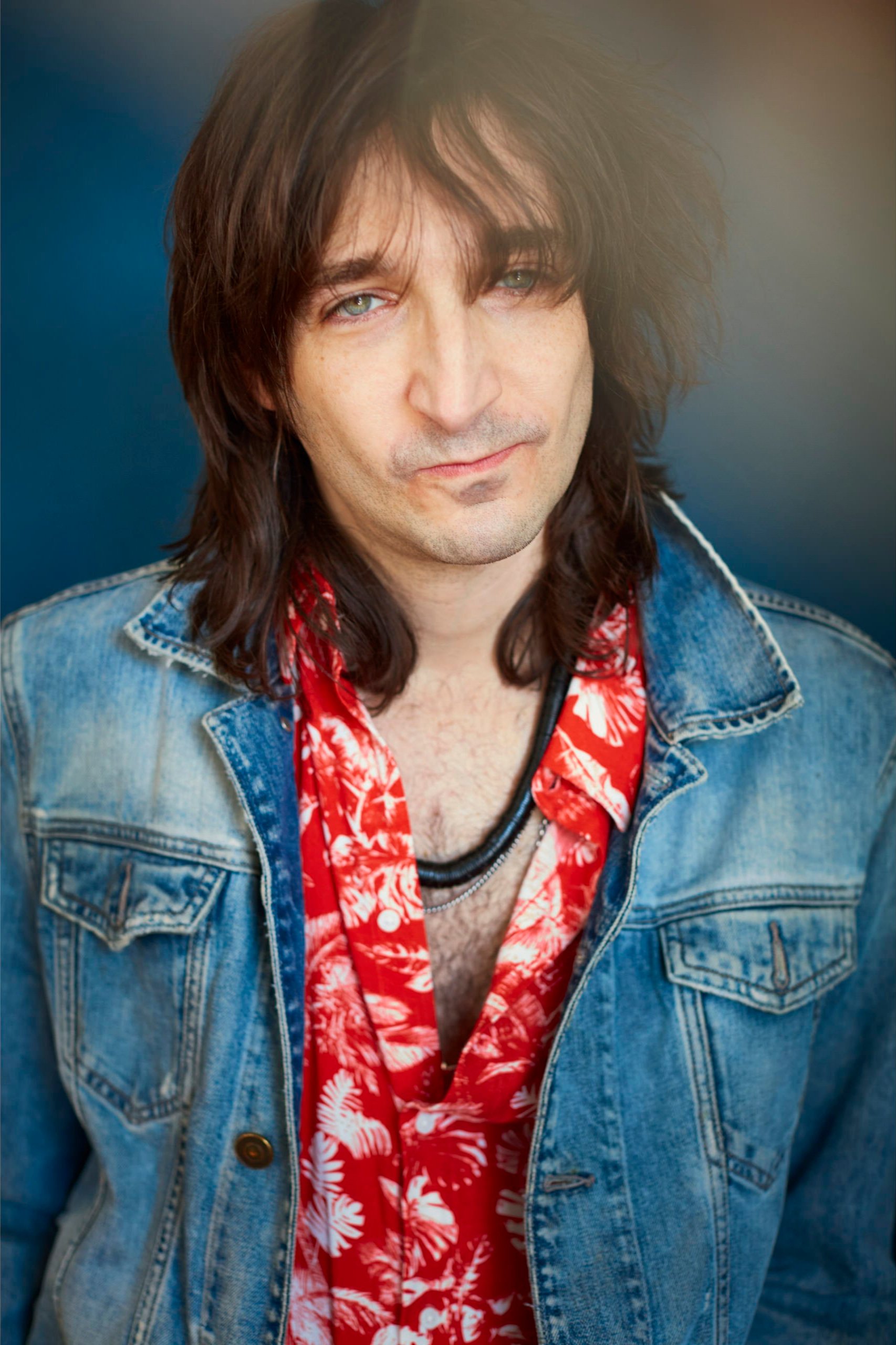 A man with long dark hair, wearing a denim jacket over a red Hawaiian shirt with white floral patterns, and layered necklaces, looking at the camera against a dark background.