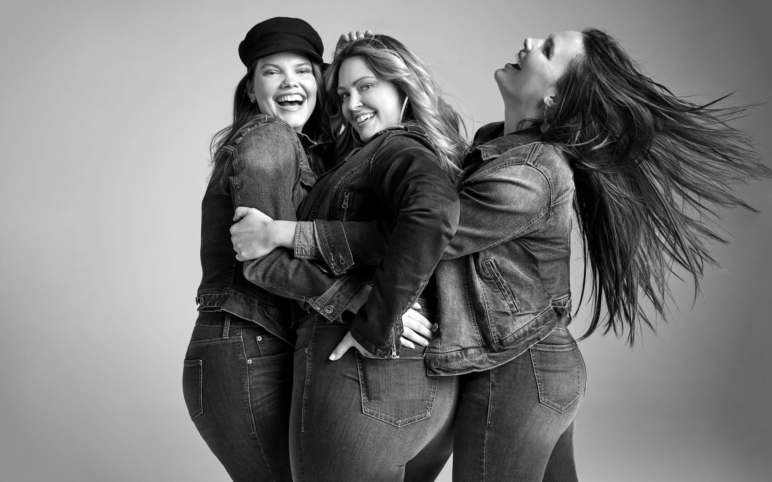 Three women wearing denim jackets and jeans, smiling and laughing, standing together in a joyful pose.
