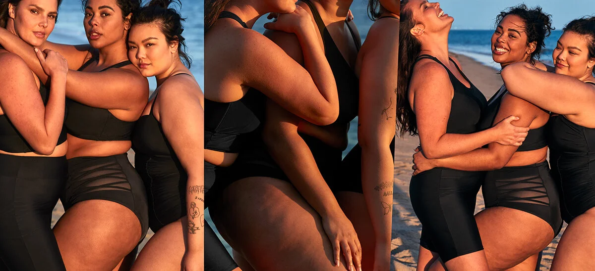 A group of women at the beach wearing black swimsuits, embracing each other and smiling, with ocean waves in the background.
