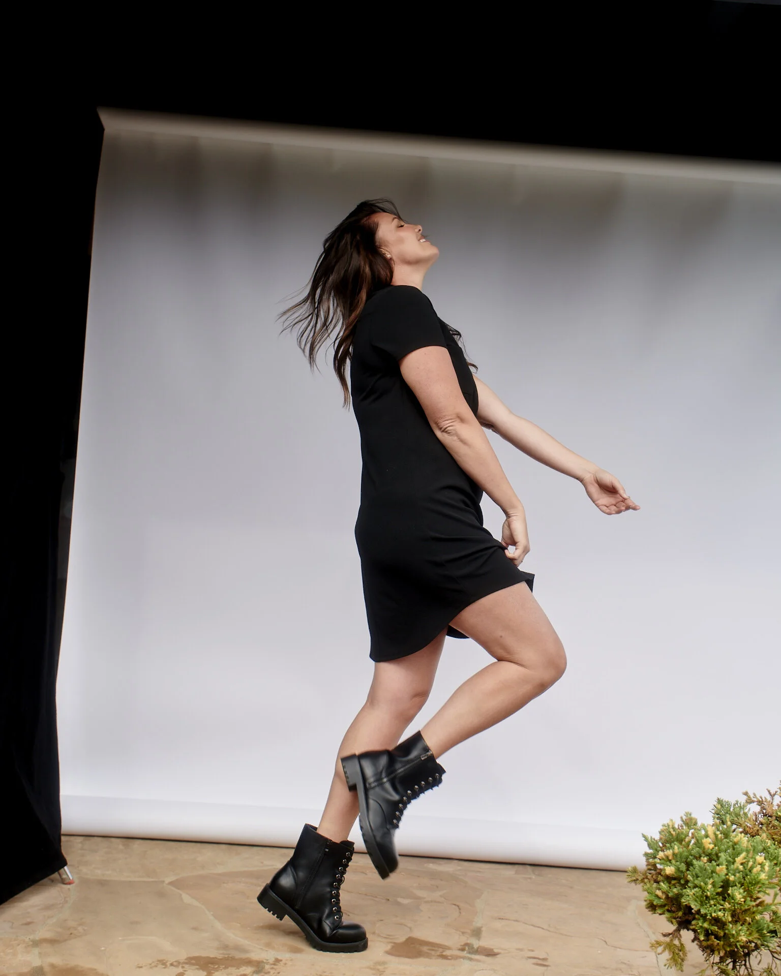 Woman wearing a black dress and black combat boots dancing or posing in front of a white backdrop.