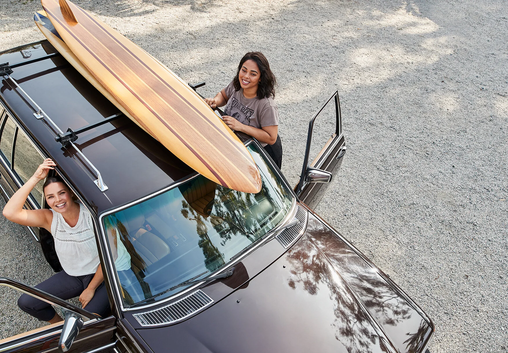 Two women with a vintage black car and a wooden surfboard on top. One woman is sitting inside the car, smiling and looking up. The other woman is standing outside with the driver's side door open and smiling, with her hand on the door.