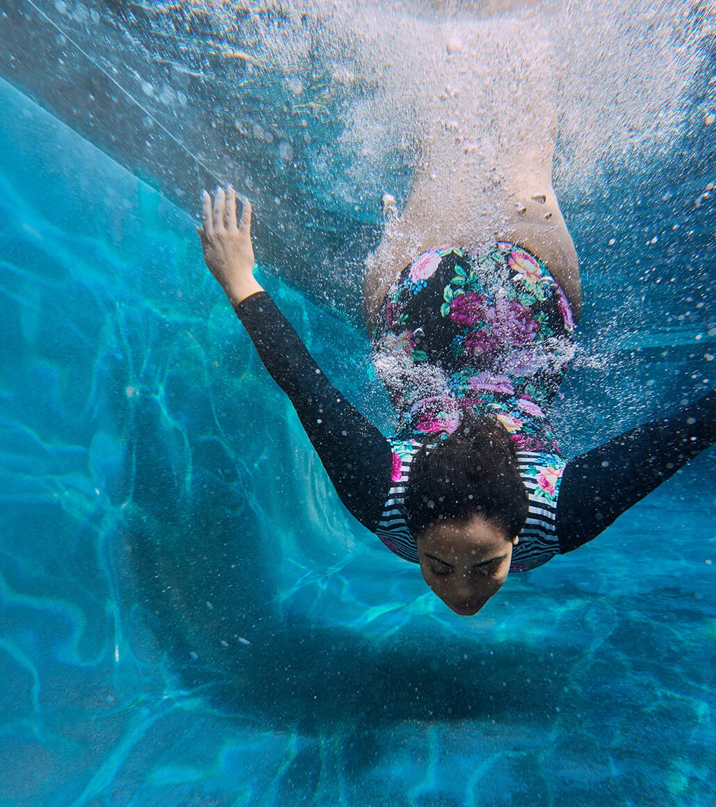 Woman swimming underwater in a pool, wearing a floral swimsuit and black sleeves.