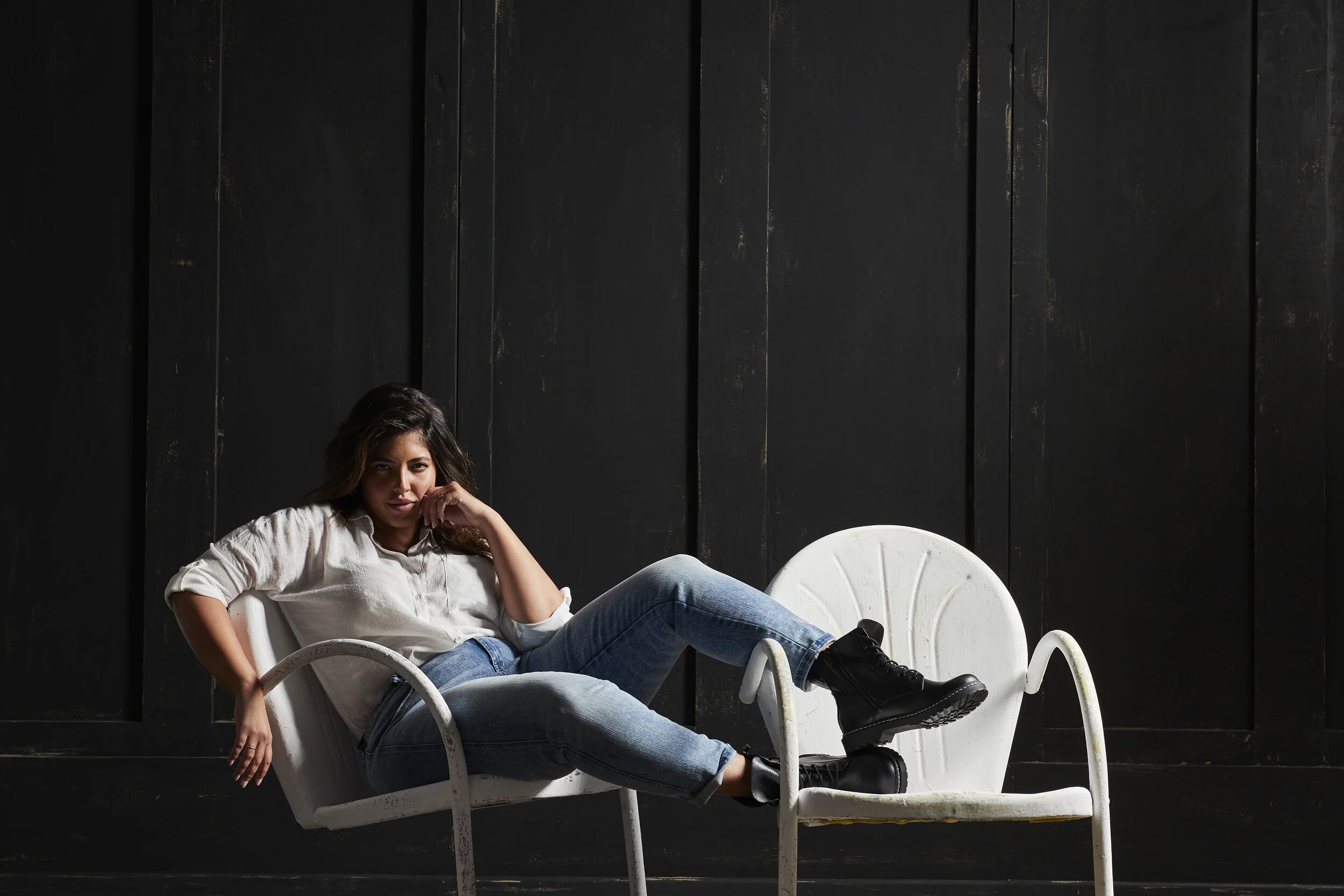 Woman lying on a white metal chair with black boots, sitting in front of a black wooden wall.