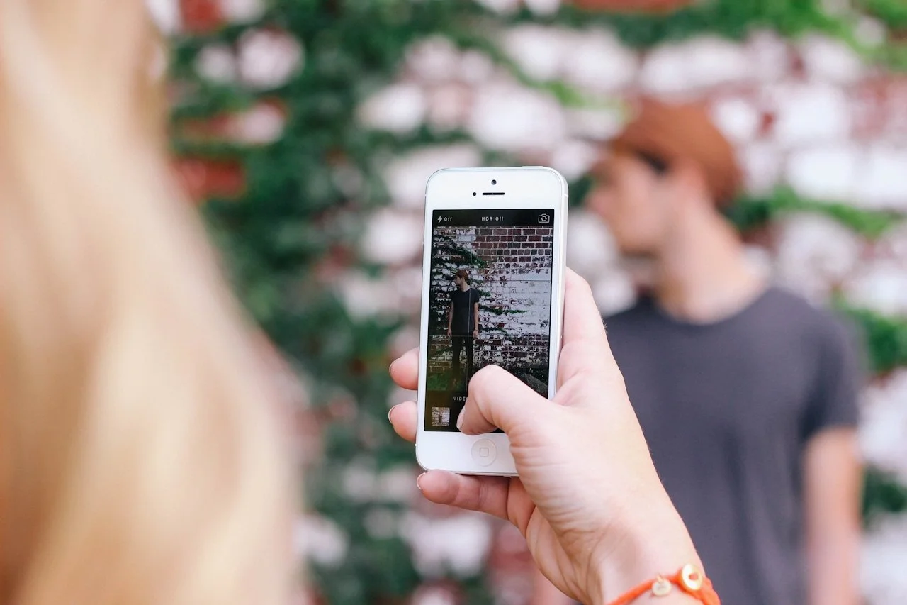 A person taking a photo of a woman with a smartphone in front of a brick and vine background.