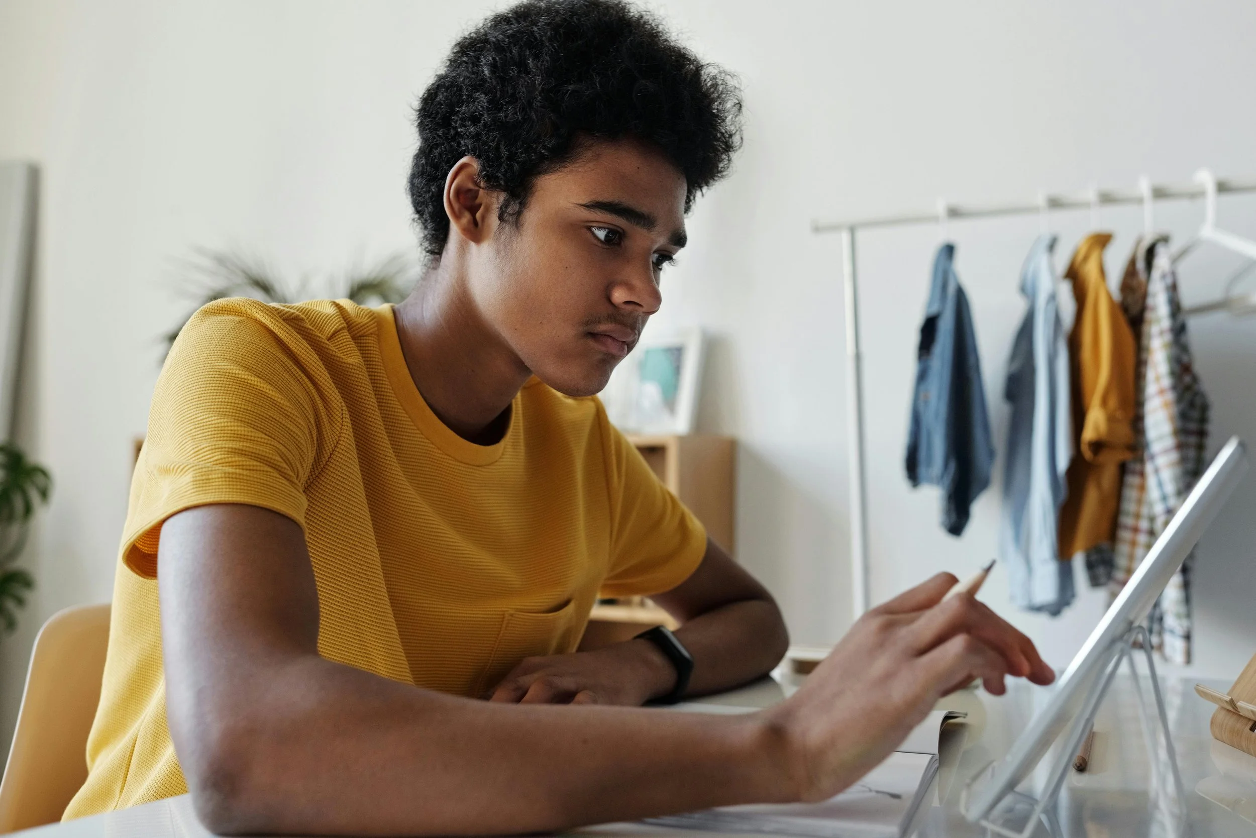 Young person in a yellow shirt using a tablet at a desk, clothing rack in the background.