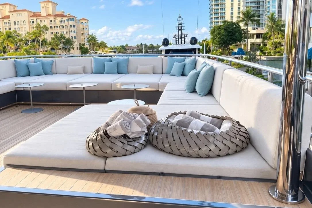 Luxury yacht deck with white seating cushions, blue and white throw pillows, two woven baskets with rolled towels, and small round tables. City high-rise buildings and palm trees are visible in the background beyond a waterway.