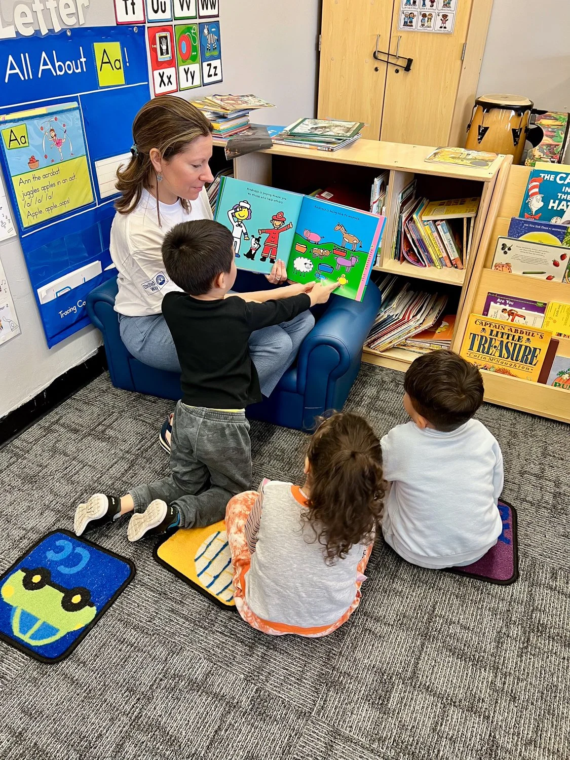 A woman with glasses and earrings sitting on a small wooden stool, reading a colorful children's book with a girl and a pizza illustration to a small group in a classroom. The background shows a large calendar for April 2023 with photos and weather info, and an alphabet wall with pictures of objects starting with each letter.