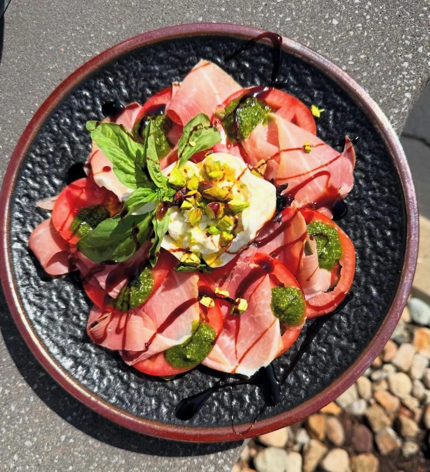 Co-Pilot Caprese- Sliced tomatoes layered with prosciutto and pistachio pesto, topped with a creamy whole burrata, crunchy pistachios, a touch of extra virgin olive oil, and a finish of aged balsamic reduction