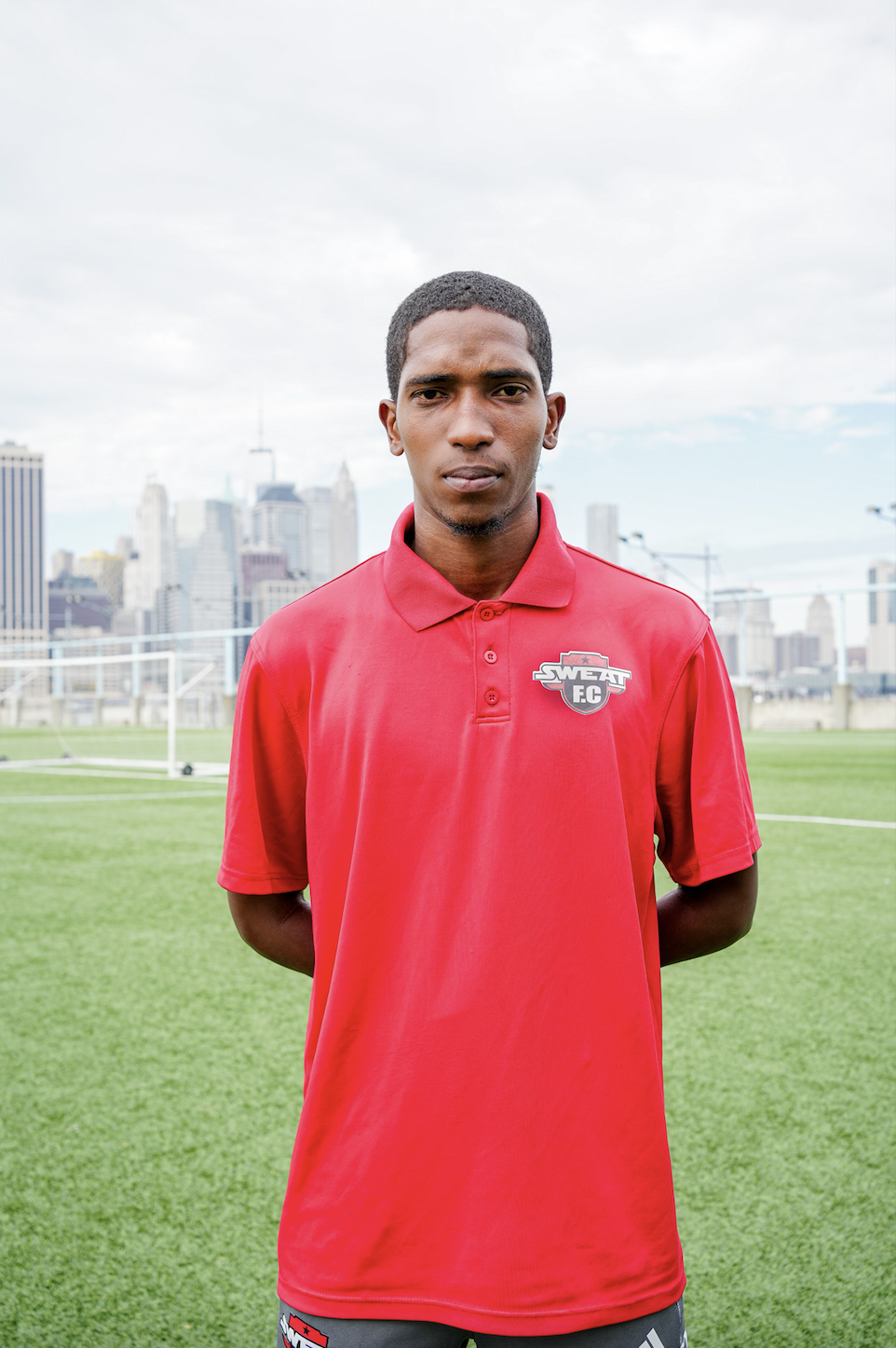 A young man standing on a soccer field in front of a city skyline, wearing a red polo shirt with a Sweat FC logo.