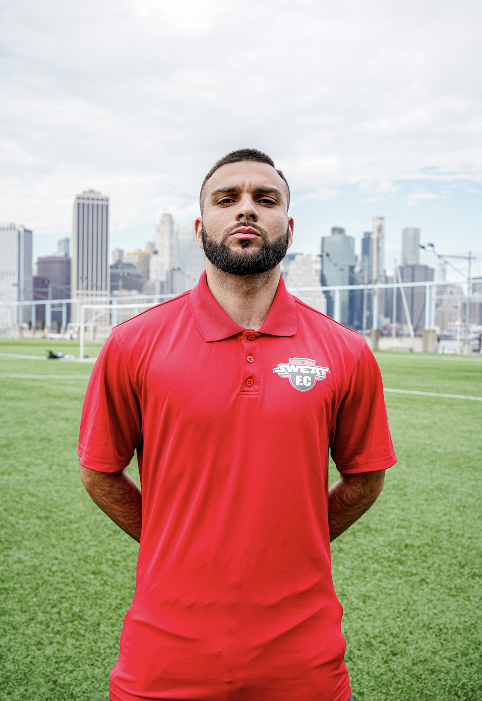 A man with a beard wearing a red polo shirt with a 'SWAT FC' logo, standing on a soccer field with a city skyline in the background.