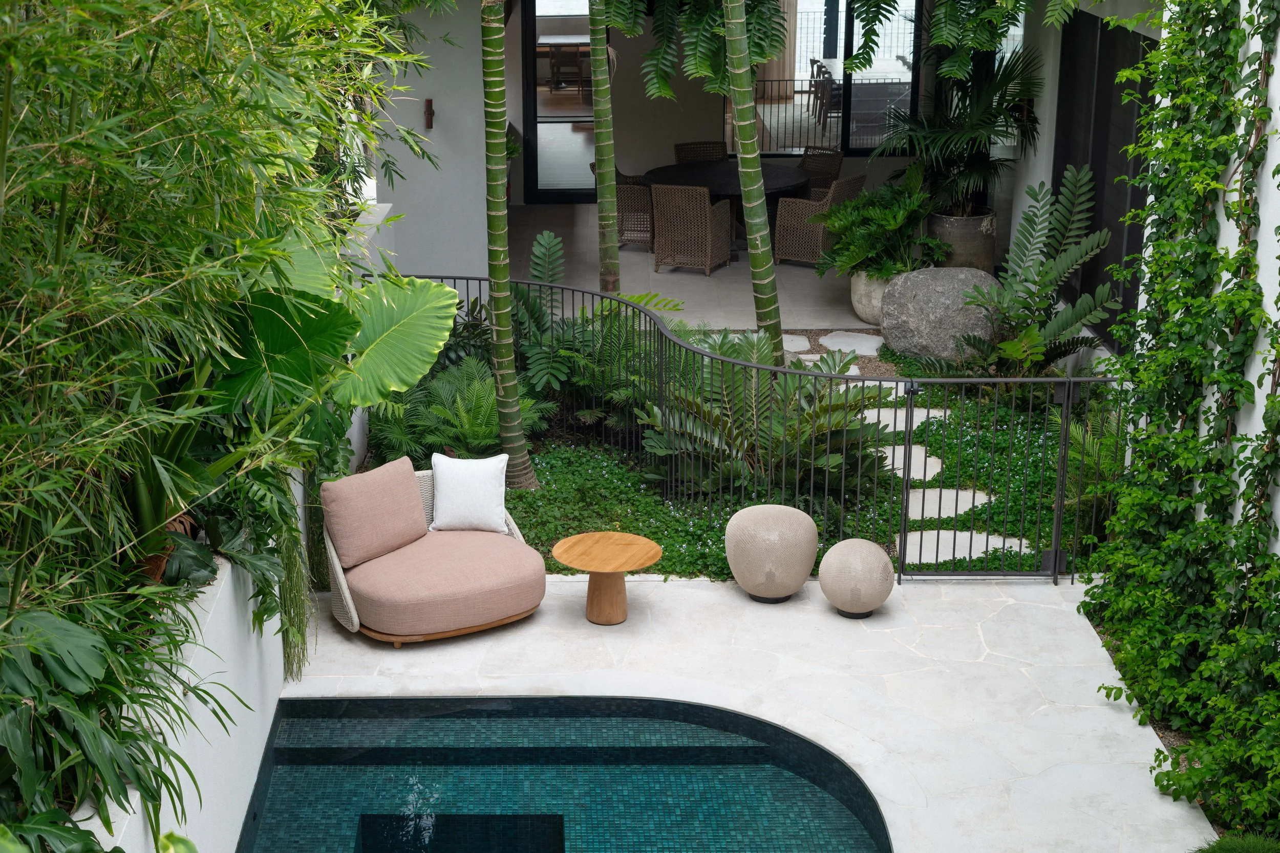 Indoor courtyard with lush green plants, a round wicker table with four chairs, and a pink cushioned outdoor sofa with pillows, overlooking a small tiled pool.