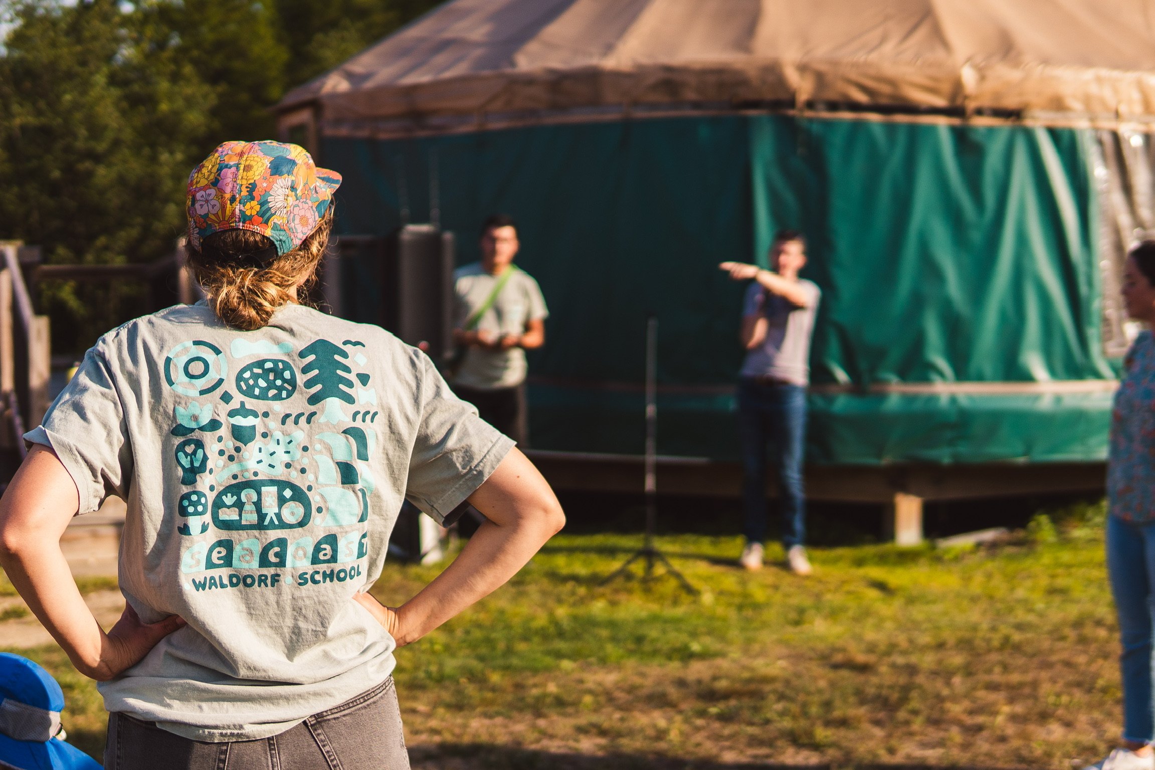 Person wearing a gray t-shirt and floral cap watching a presentation outdoors. In the background, two people are speaking or instructing in front of a green tent.