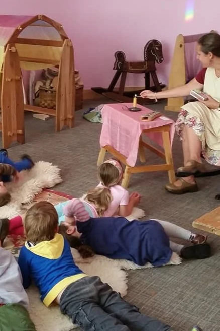 Children lying on the carpet and listening to a woman reading a story in a cozy room with wooden furniture and rocking horse toys.