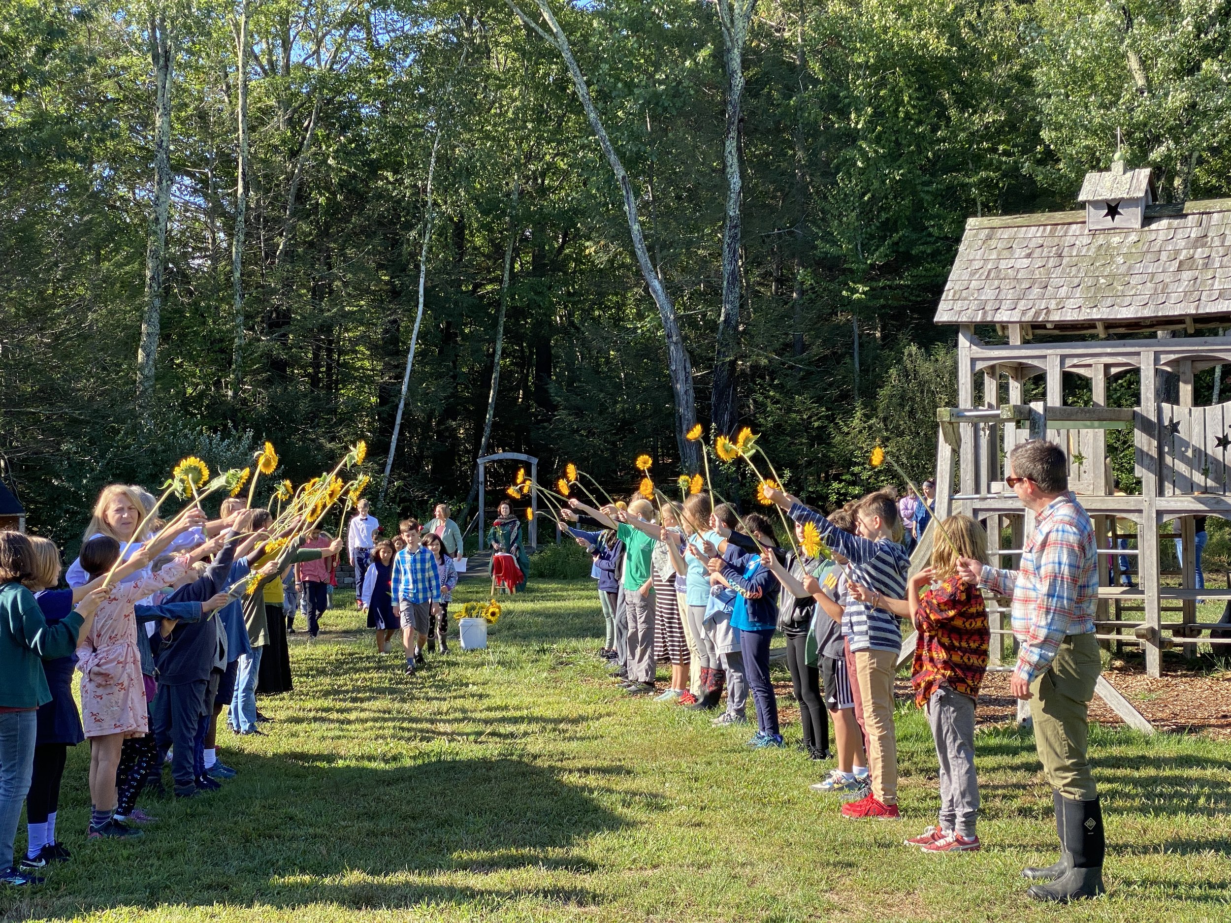 Children and adults are standing in two lines facing each other, holding bright yellow sunflowers, engaging in a coordinated outdoor activity in a grassy area with trees and a wooden structure in the background.