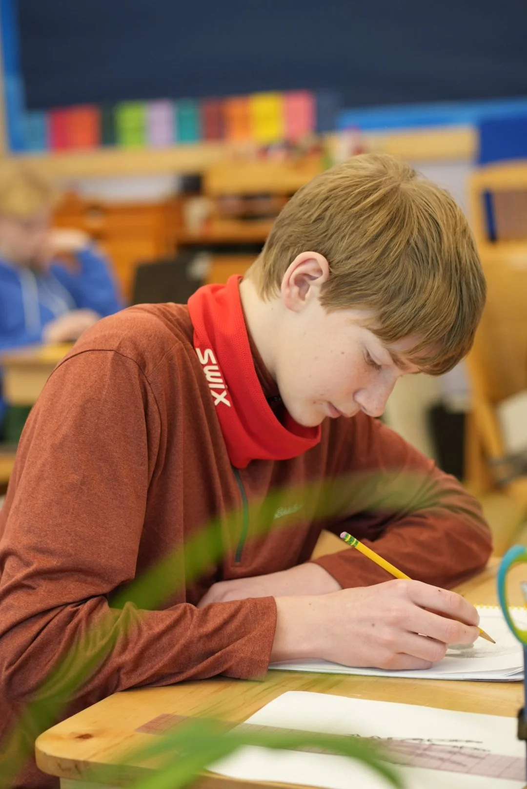 A young boy with light brown hair and freckles, wearing a red neck gaiter with white text, concentrating on writing with a yellow pencil at a wooden desk in a classroom.