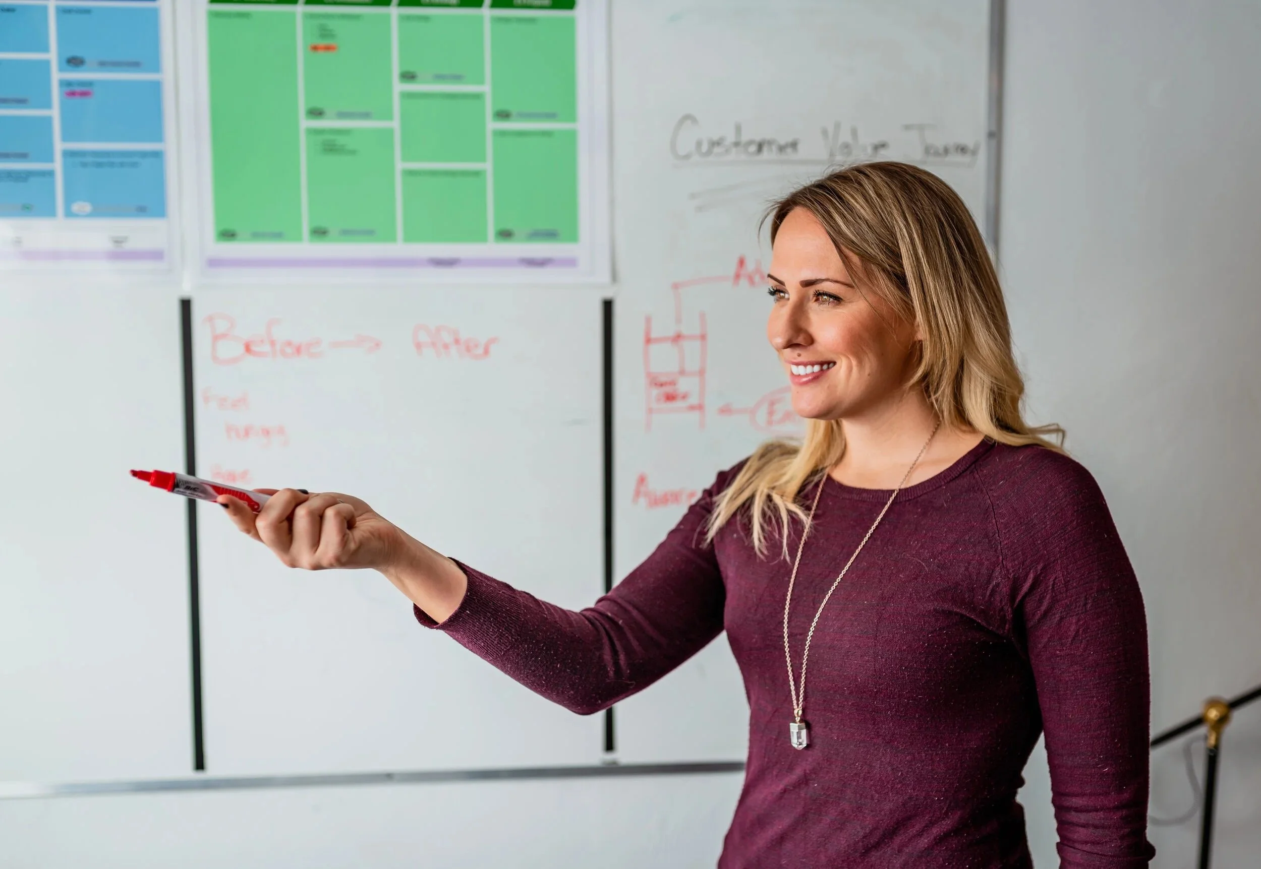 A woman smiling and holding a red marker in front of a whiteboard with colorful charts and handwritten notes.