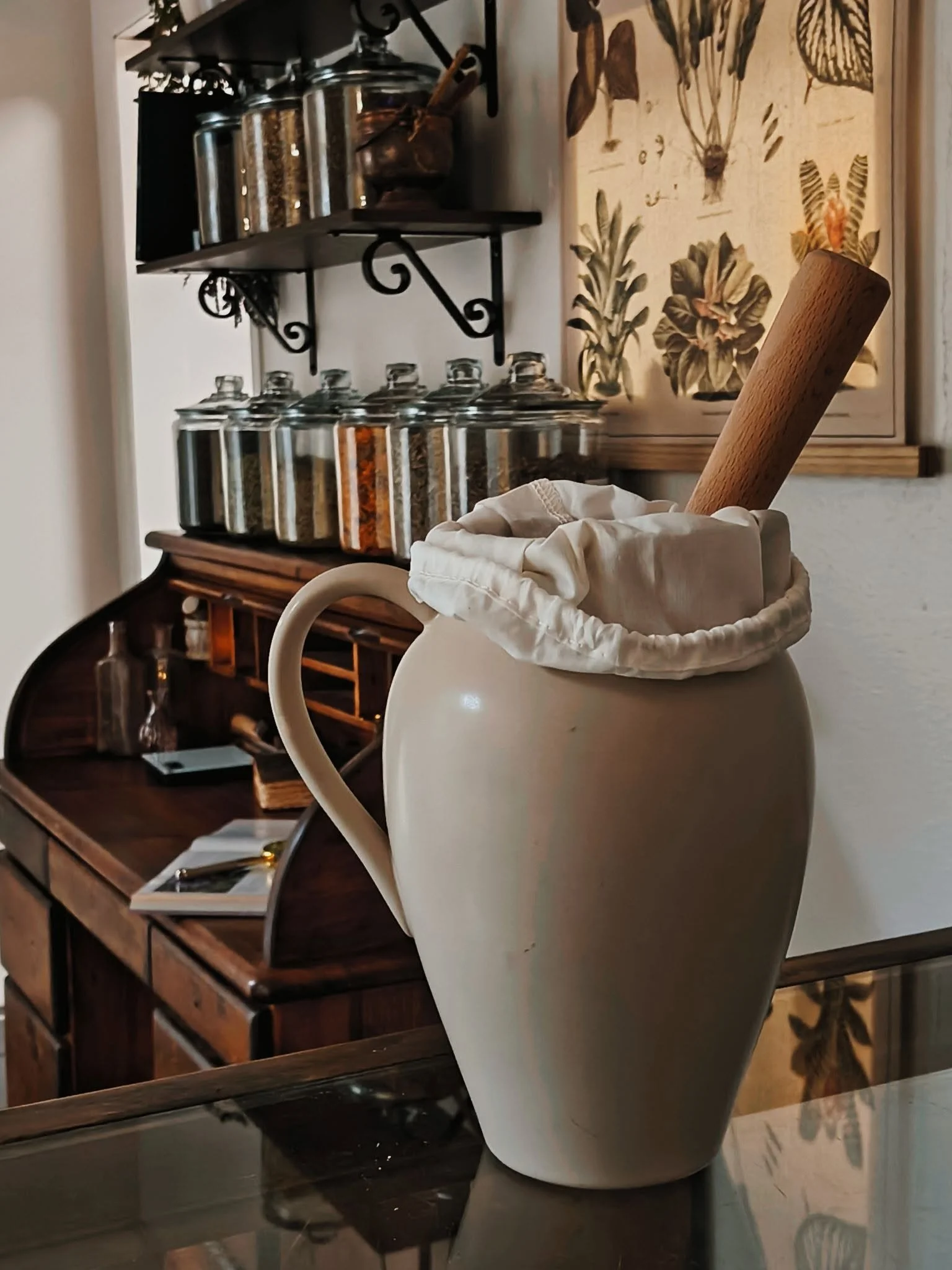 A ceramic pitcher with a wooden pestle inside it, placed on a reflective surface in front of a wooden sideboard with open shelves and white drawers. Behind, there is a wall hanging with botanical illustrations and a black metal shelf holding glass jars and small containers.
