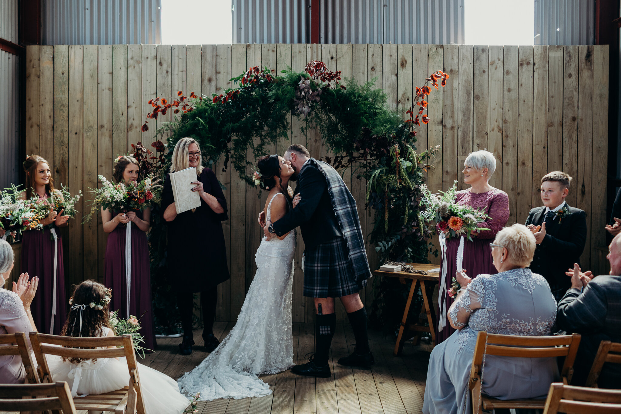 bride and groom kissing under greenery arch by sparrow &amp; rose flowers in barn outdoors at dalduff farm photographed by wedding photographer ayrshire scotland