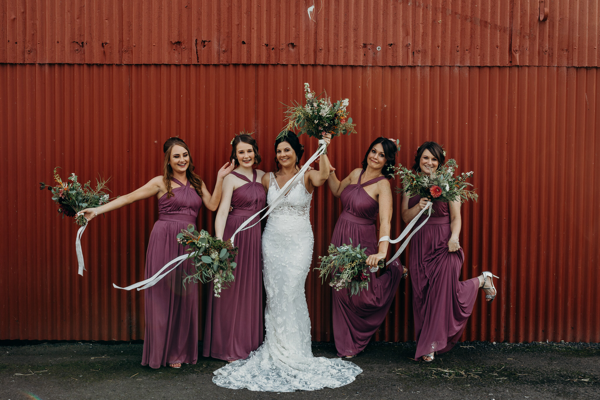 bride wearing unbridaled dress and bridesmaids standing in front of red barn holding bouquets by sparrow &amp; rose flowers at dalduff barn photographed by wedding photographer ayrshire scotland